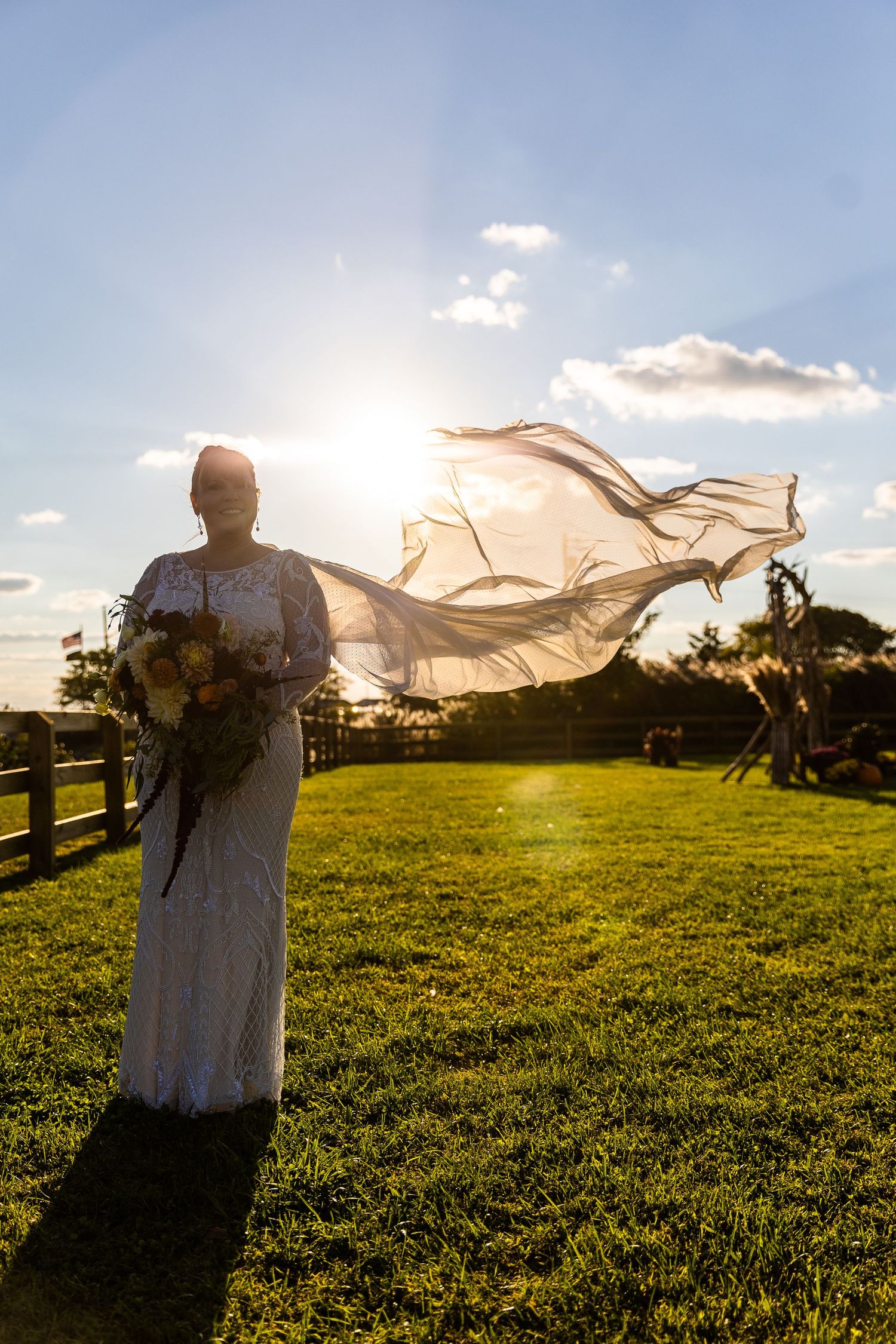 Bride in white lace gown with veil flowing in wind backlit by golden sunset on green lawn.