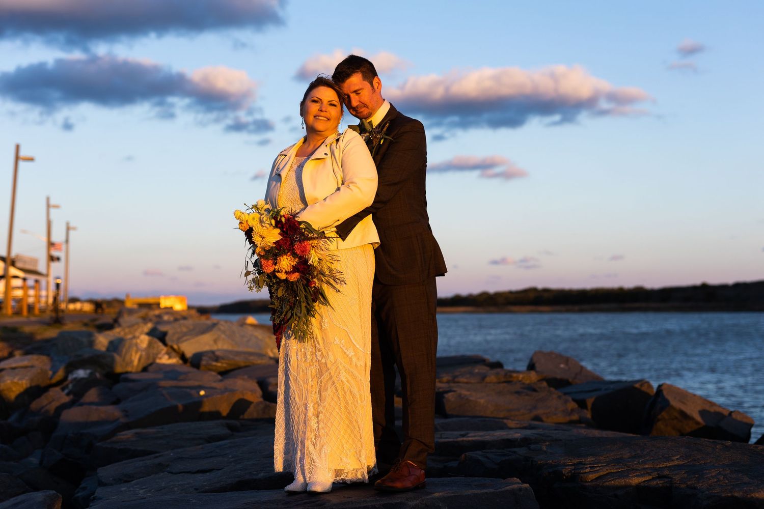 Newlyweds standing close together on coastal rocks with colorful bouquet under blue twilight sky.