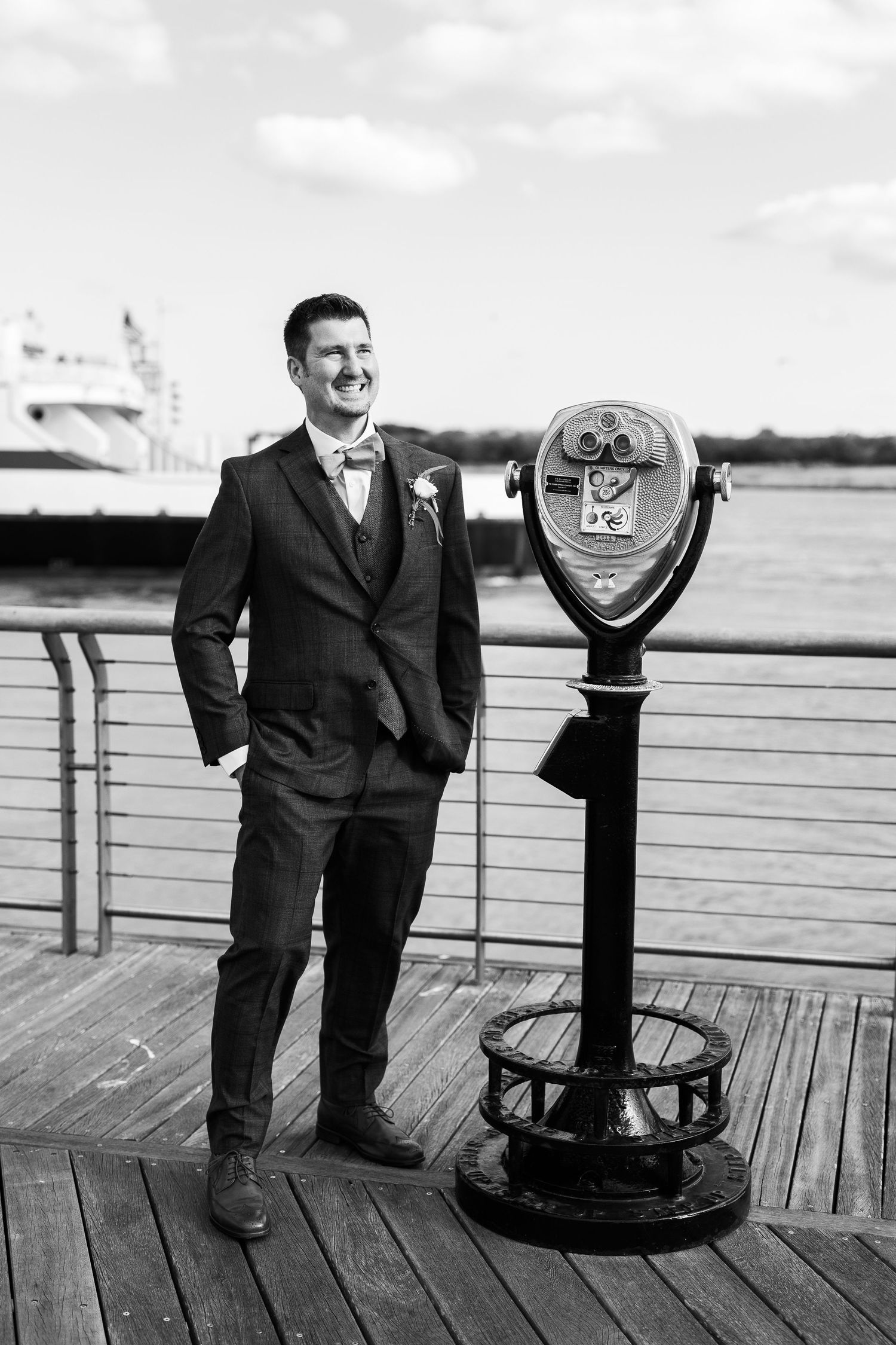 Smiling groom in dark suit standing beside coin-operated binoculars on waterfront boardwalk.