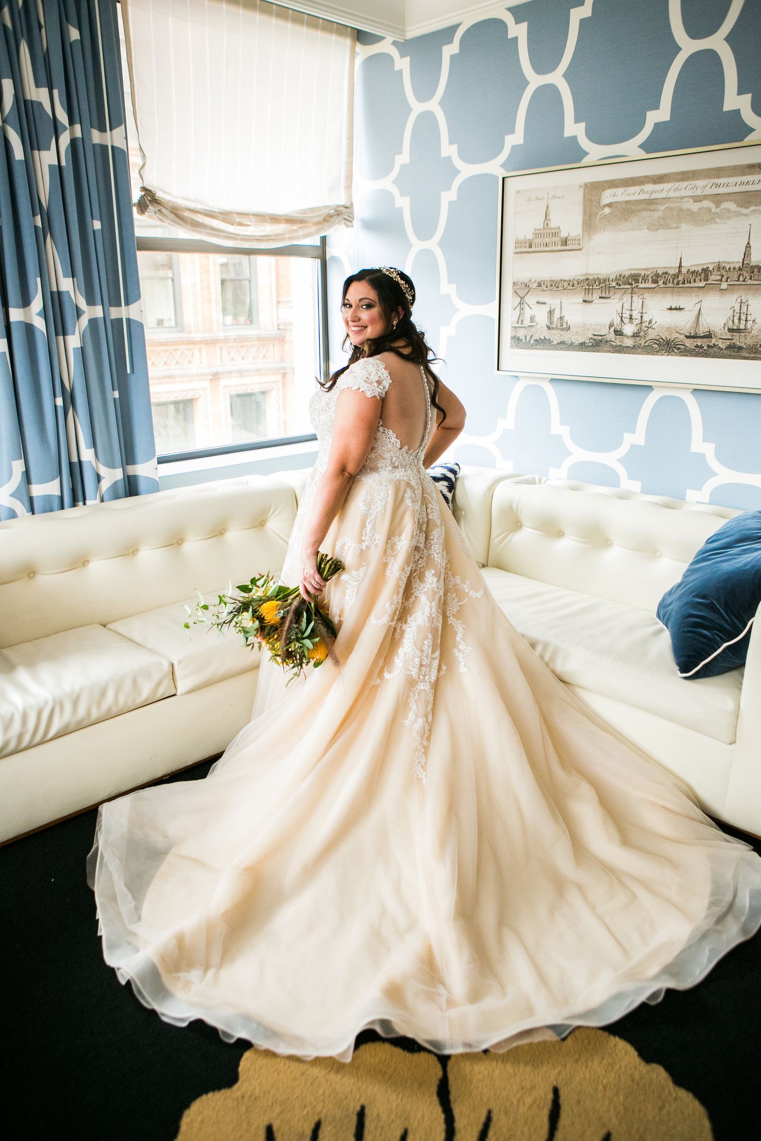 Bride in a stunning champagne lace ball gown with long train holds a colorful bouquet by a window.