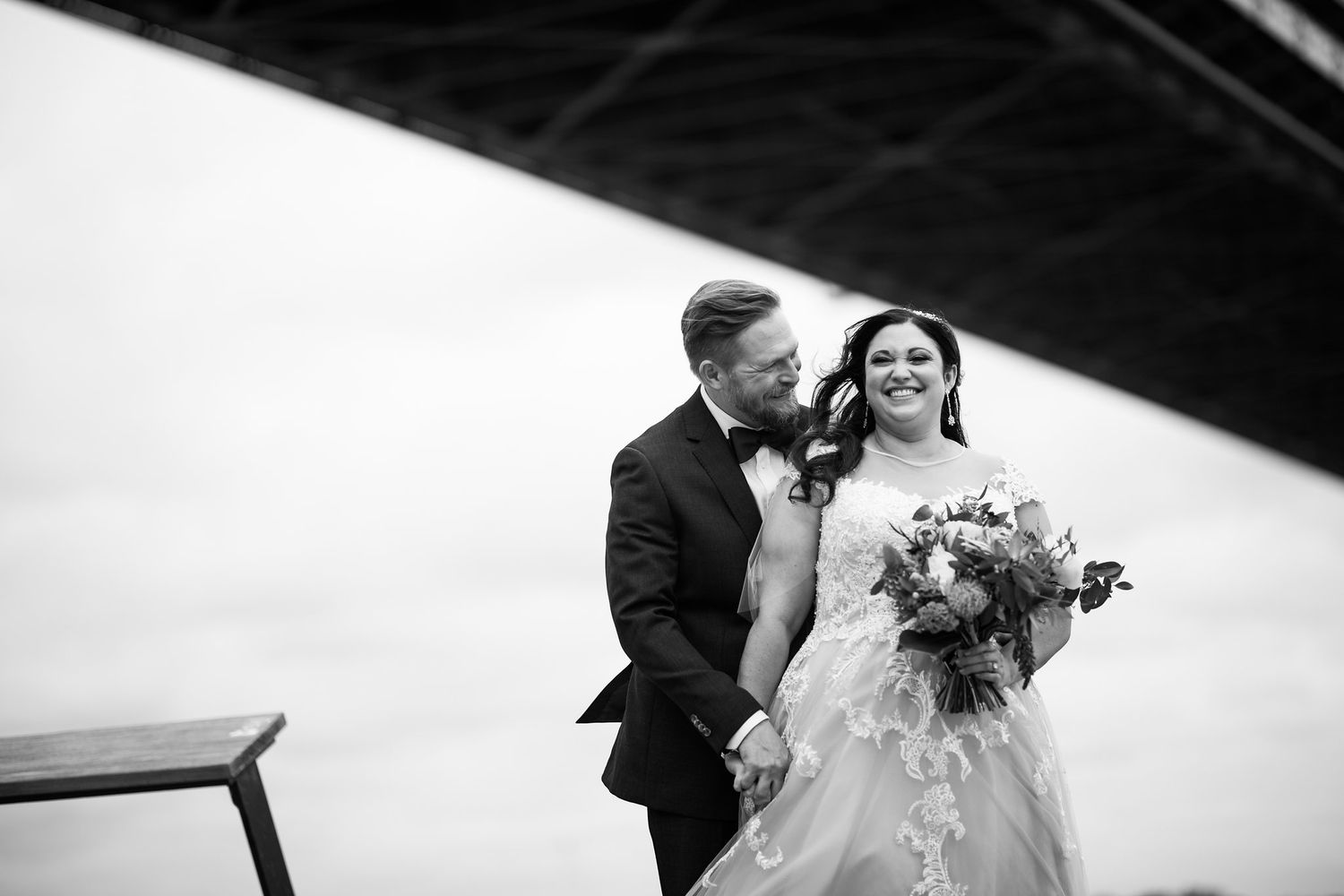 Black and white photo of groom whispering to laughing bride near a bridge, bride holding a bouquet.