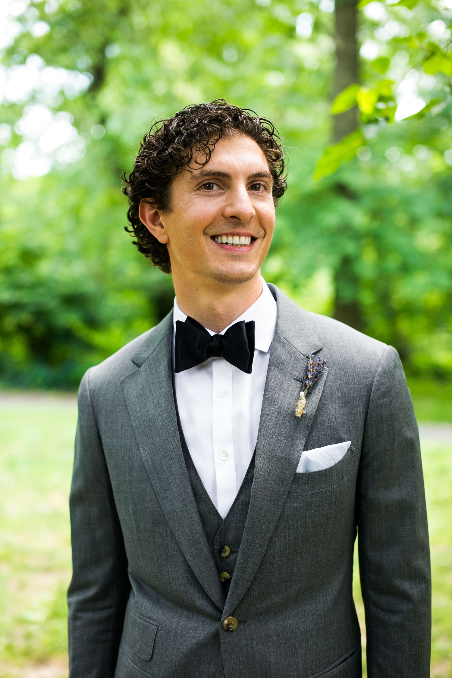 Handsome groom with curly hair smiles outdoors wearing gray three-piece suit with black bow tie and boutonniere.