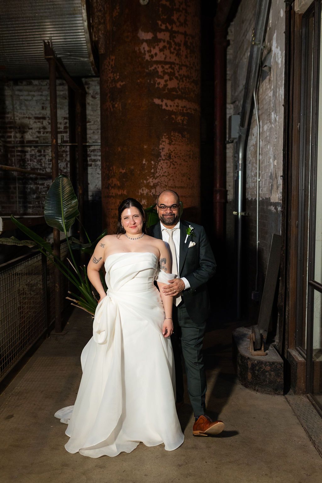 Elegant bride and groom posing together in rustic industrial wedding venue with brick walls.
