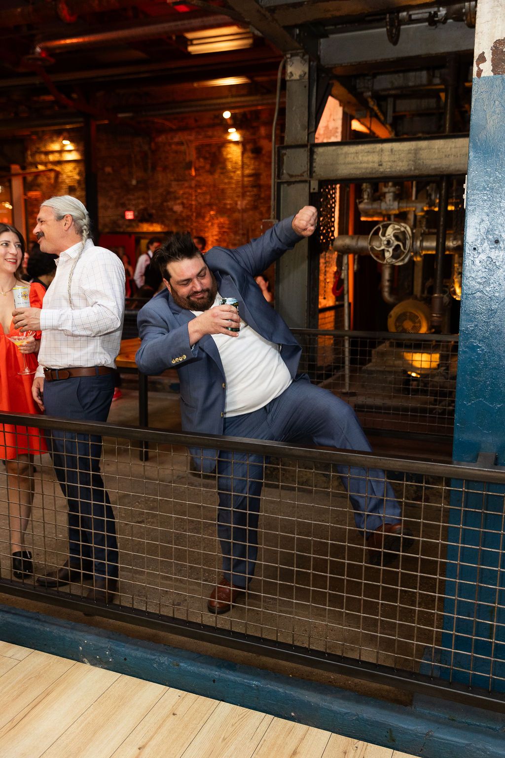A man in a blue suit jumps energetically at a lively indoor party in an industrial space.