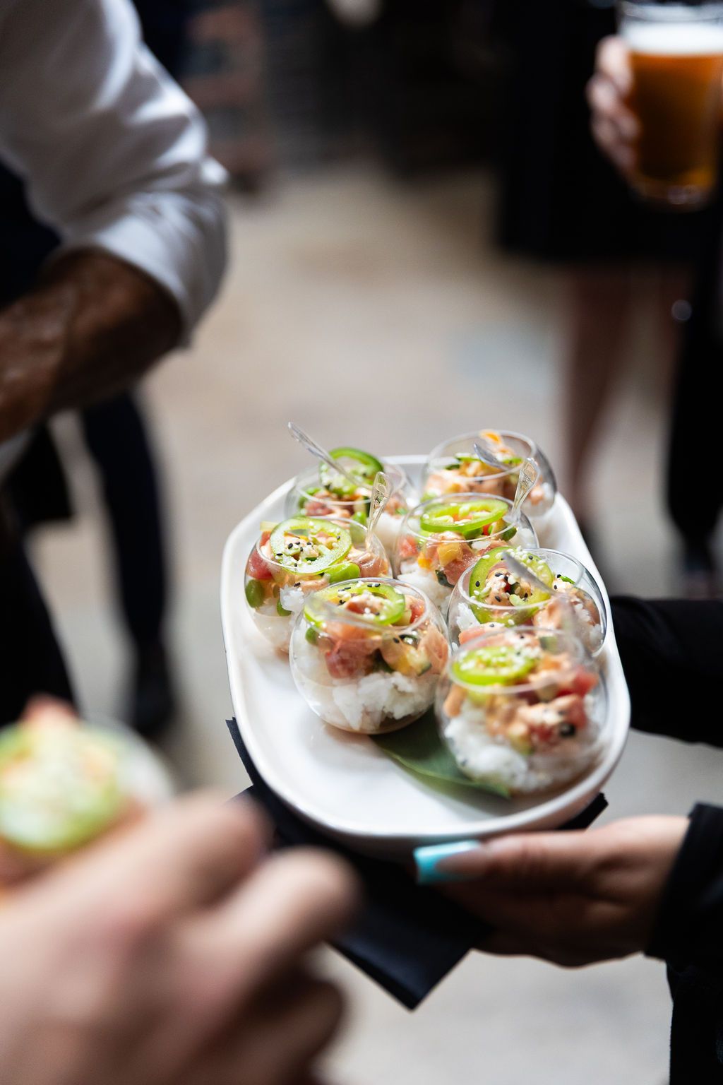Server holding a tray of gourmet seafood appetizers in glasses garnished with jalapeño at a catered event.