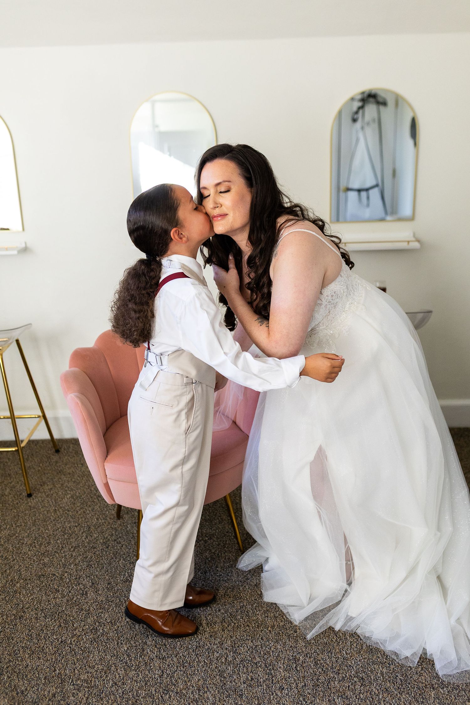 Young boy in white suit kissing bride in wedding dress on the cheek indoors.
