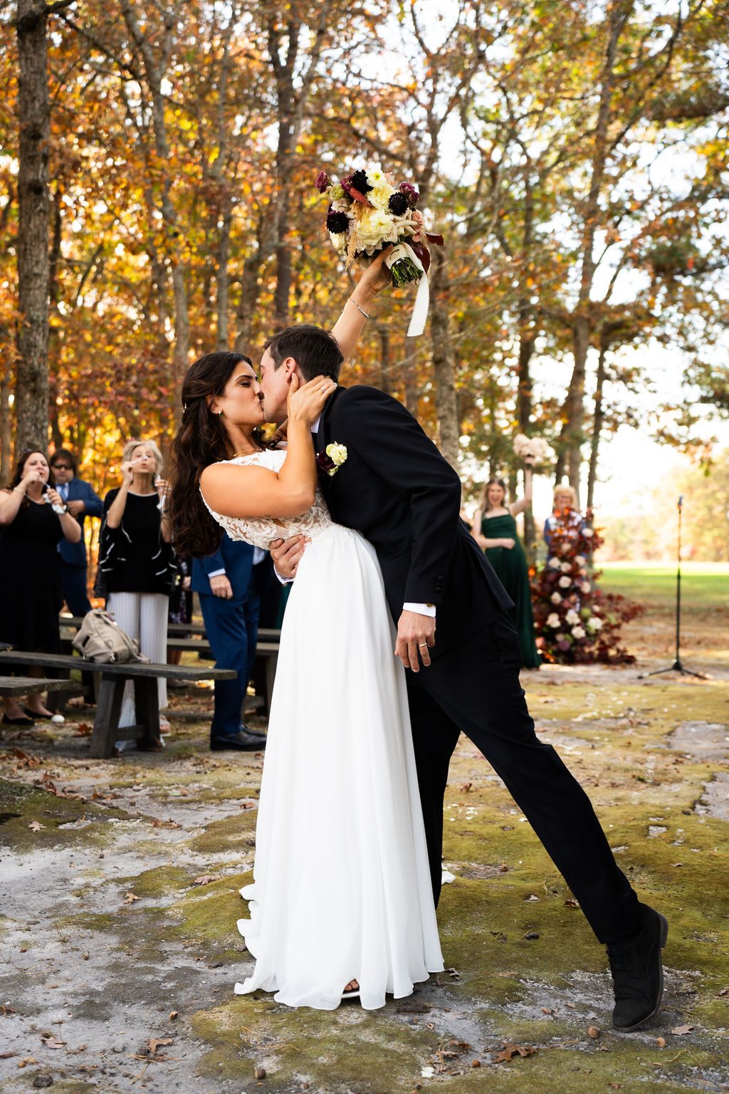 Newlyweds share romantic first kiss as bride raises bouquet amid cheering guests at fall outdoor wedding.