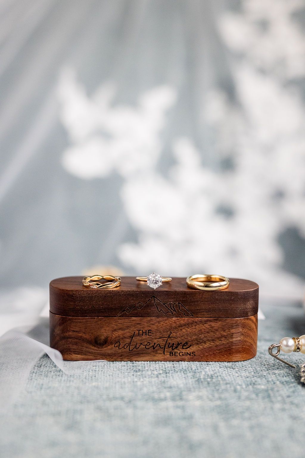 Three wedding rings displayed on a wooden adventure begins ring box with a lace veil in the background.