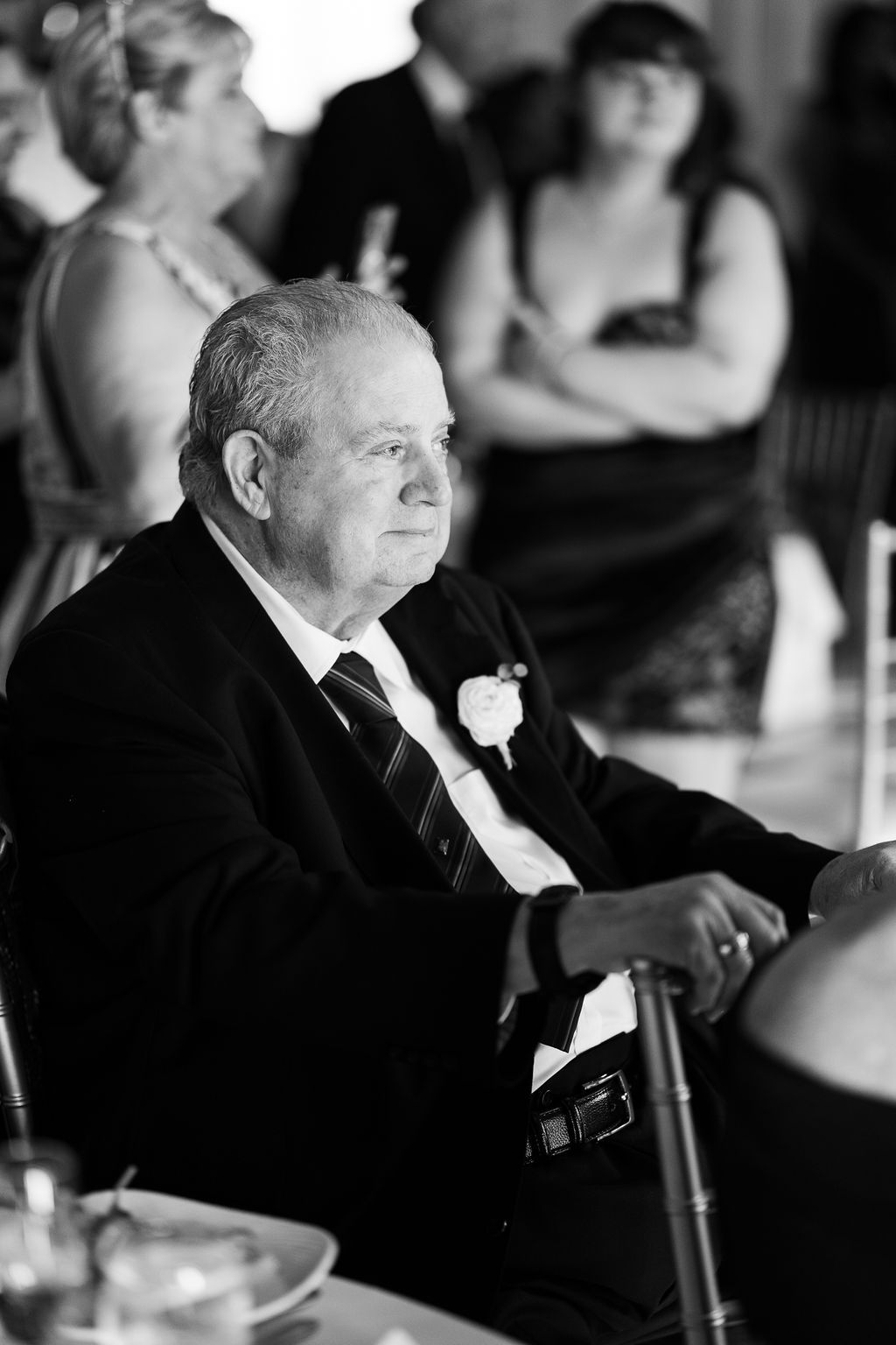 Elderly wedding guest in black suit with boutonniere seated at reception table, black and white photo.