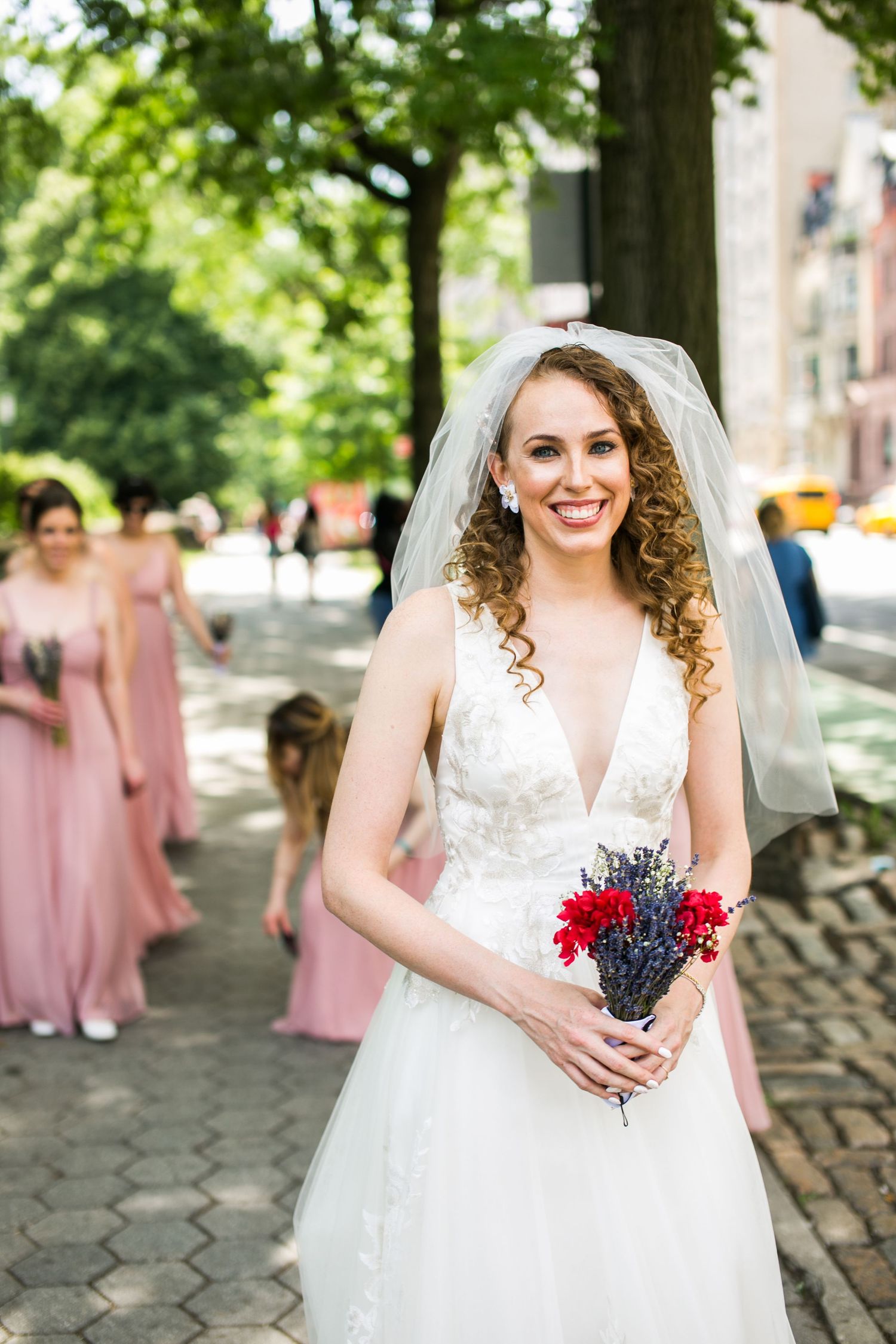Radiant bride in white lace dress and veil smiles while holding red floral bouquet outdoors.