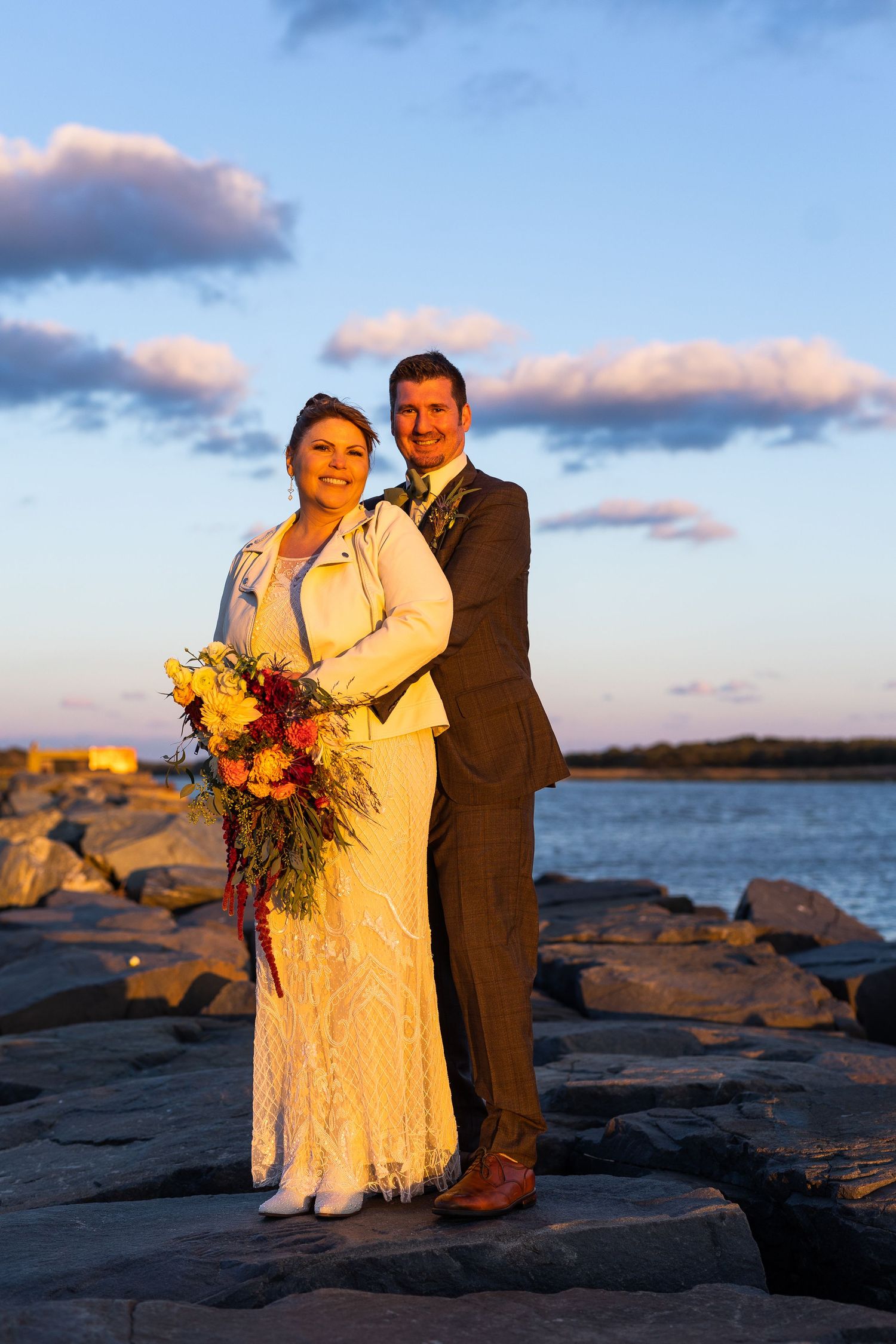 Bride and groom posing on rocky waterfront at golden hour with vibrant autumn wedding bouquet.