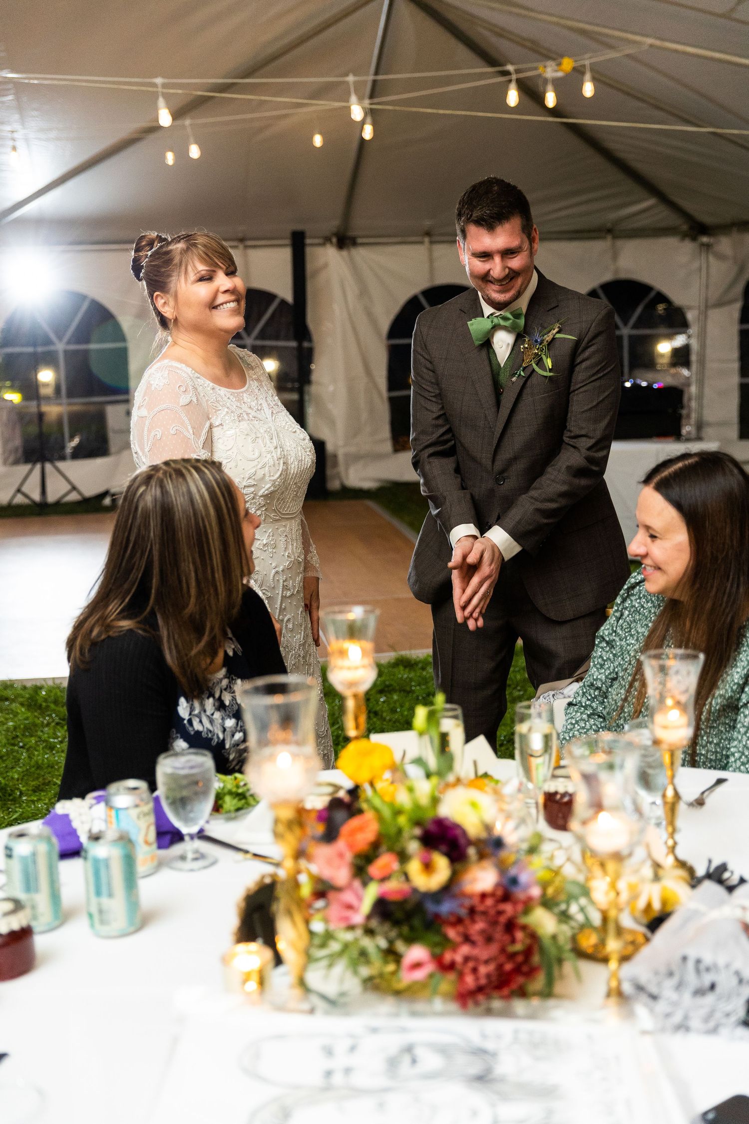 Bride and groom greeting seated guests at candlelit wedding reception table with colorful florals.
