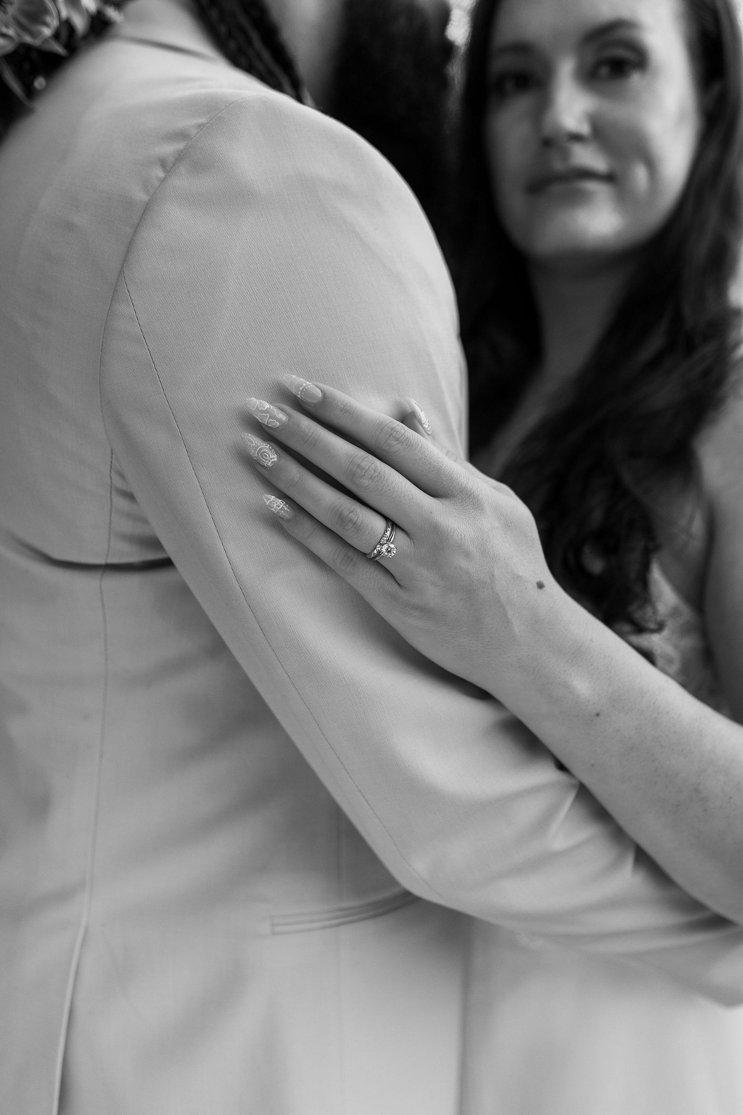 Close-up black and white detail of bride's hand with engagement ring resting on groom's suit jacket.