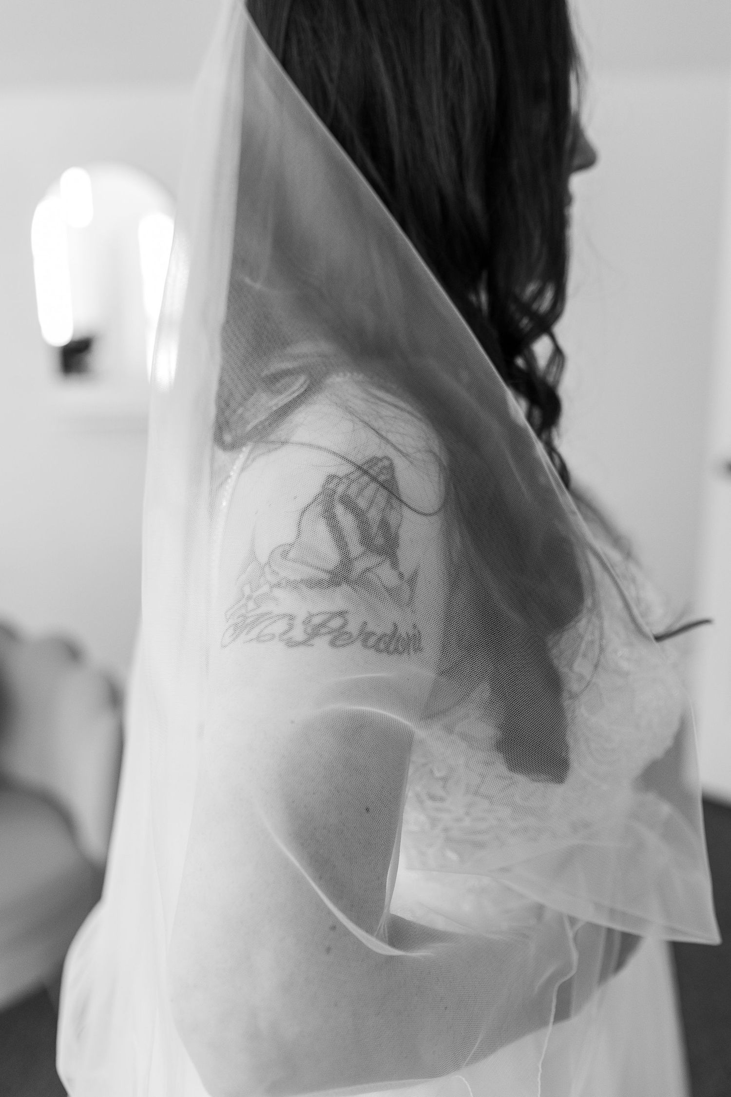 Black and white close-up of bride's shoulder tattoo beneath sheer wedding veil.