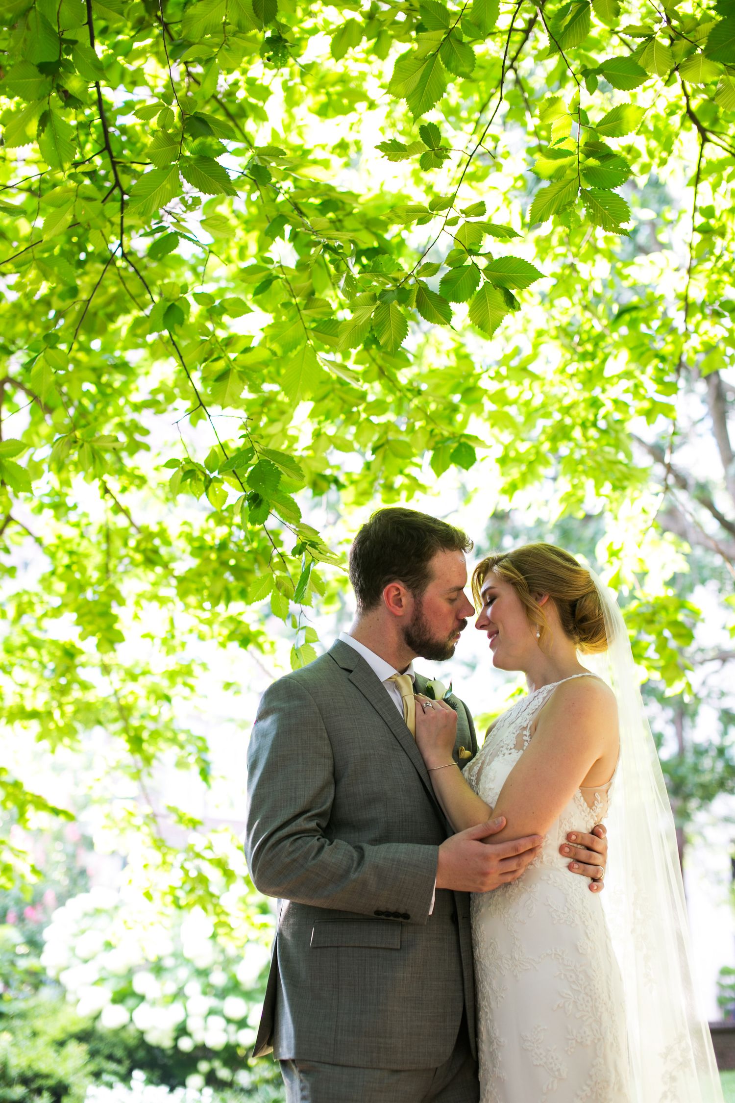 Bride and groom share a romantic almost-kiss under lush green tree branches in an outdoor garden wedding portrait.
