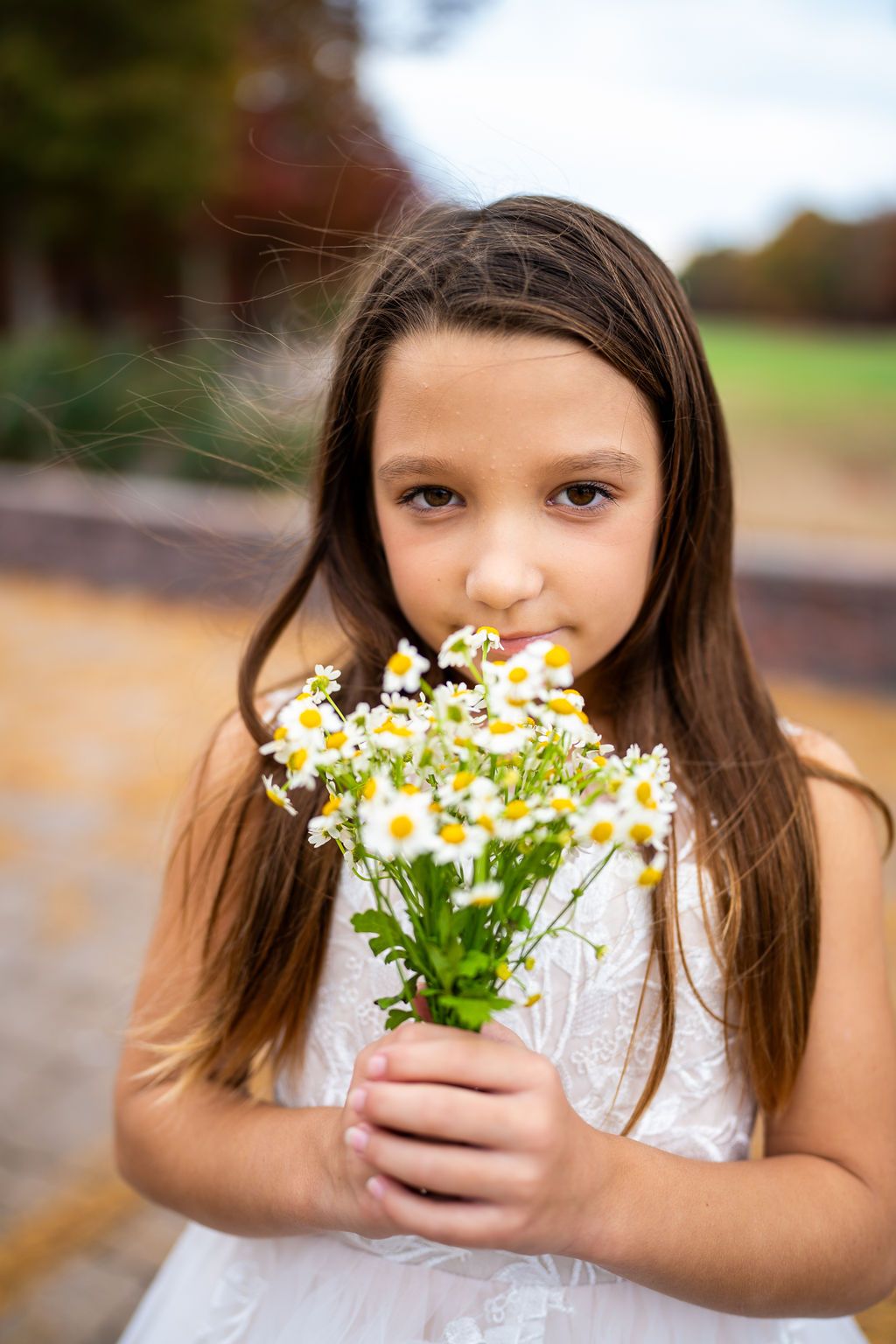 Young flower girl in white dress holds bouquet of white daisies outdoors in autumn.