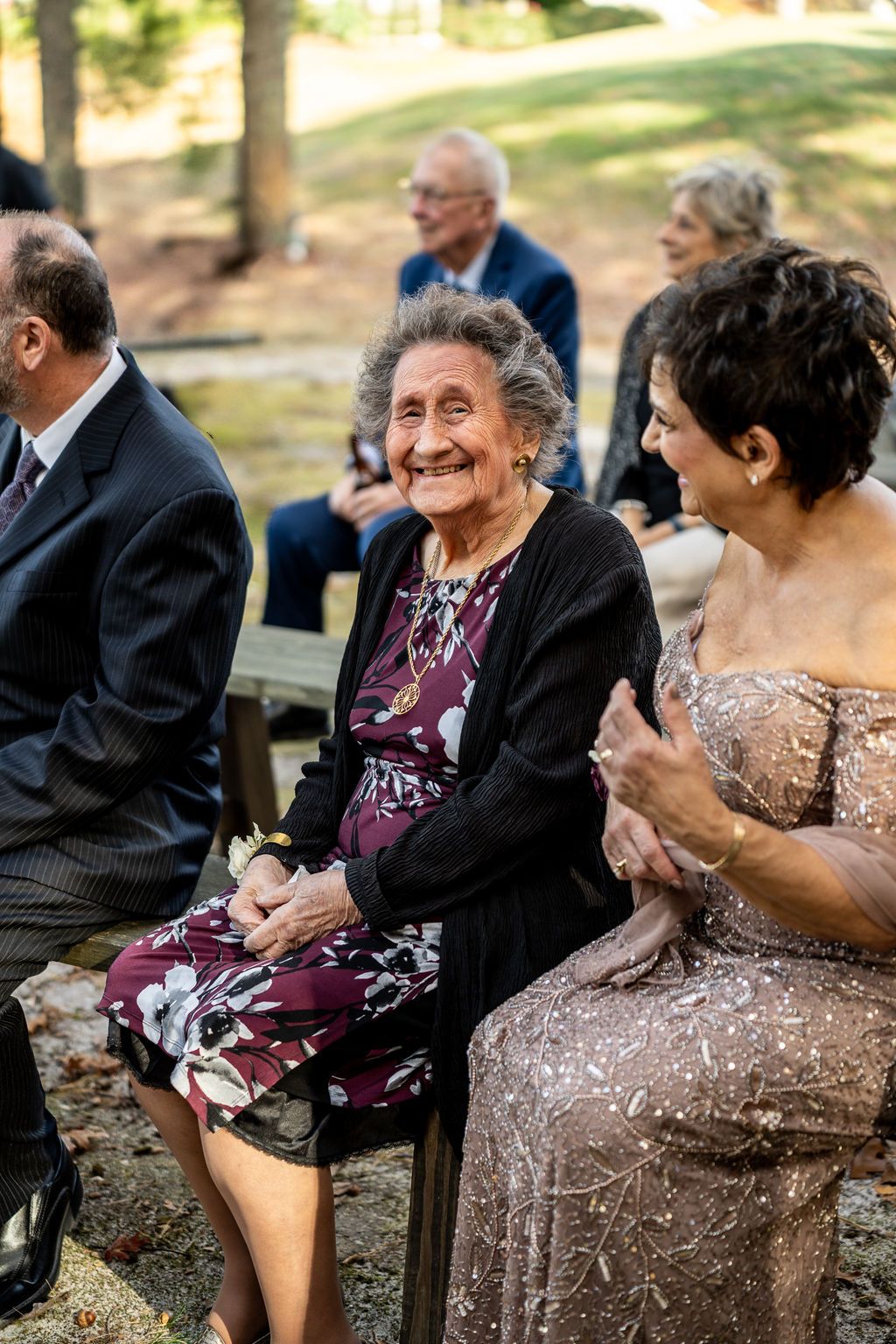 Elderly woman smiles joyfully while seated at outdoor wedding ceremony beside woman in sequined dress.