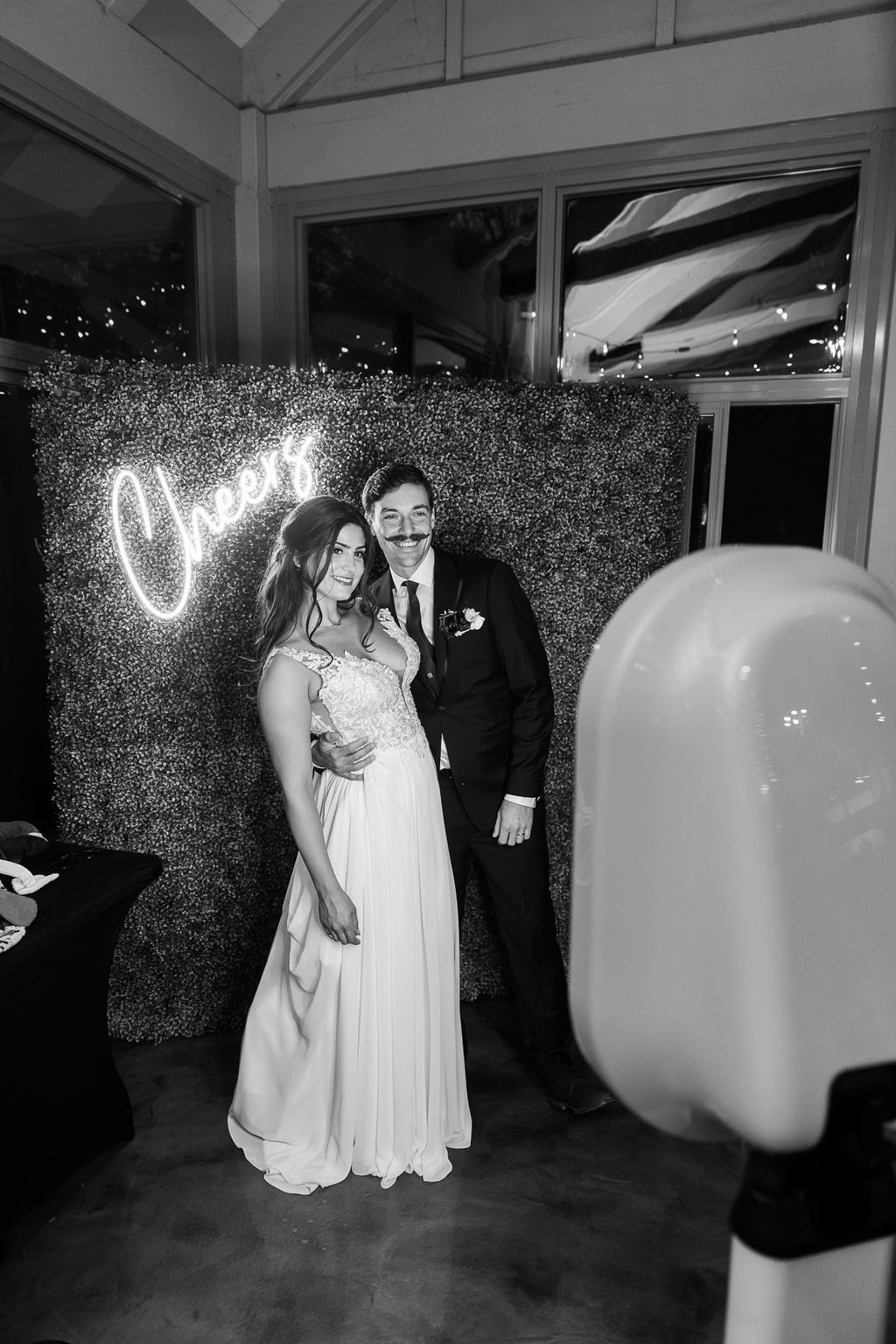 Bride and groom pose in black and white photo at a wedding photo booth with a neon Cheers sign.