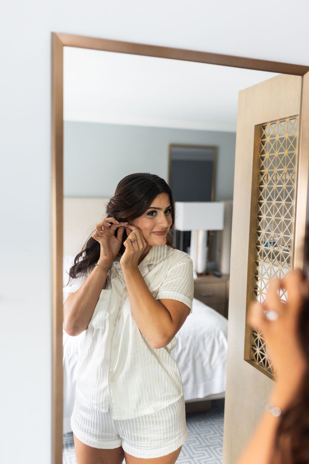 Brunette bride in striped pajamas putting on earrings while smiling at her reflection in a large mirror.