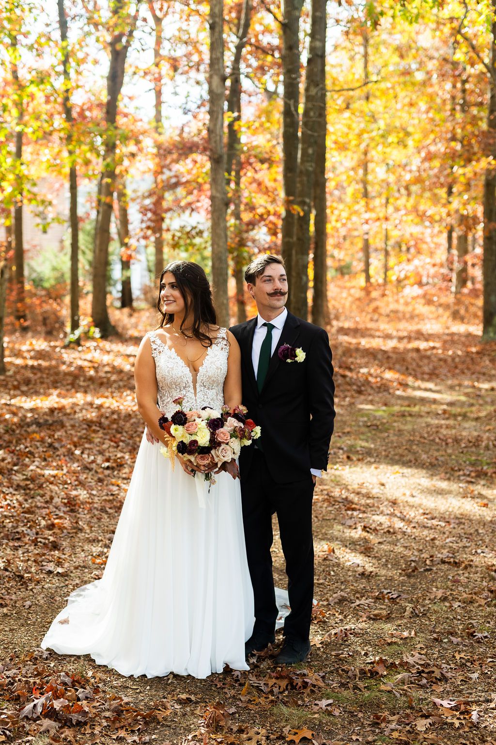 Bride holding burgundy bouquet standing with groom in black suit amid colorful autumn forest backdrop.