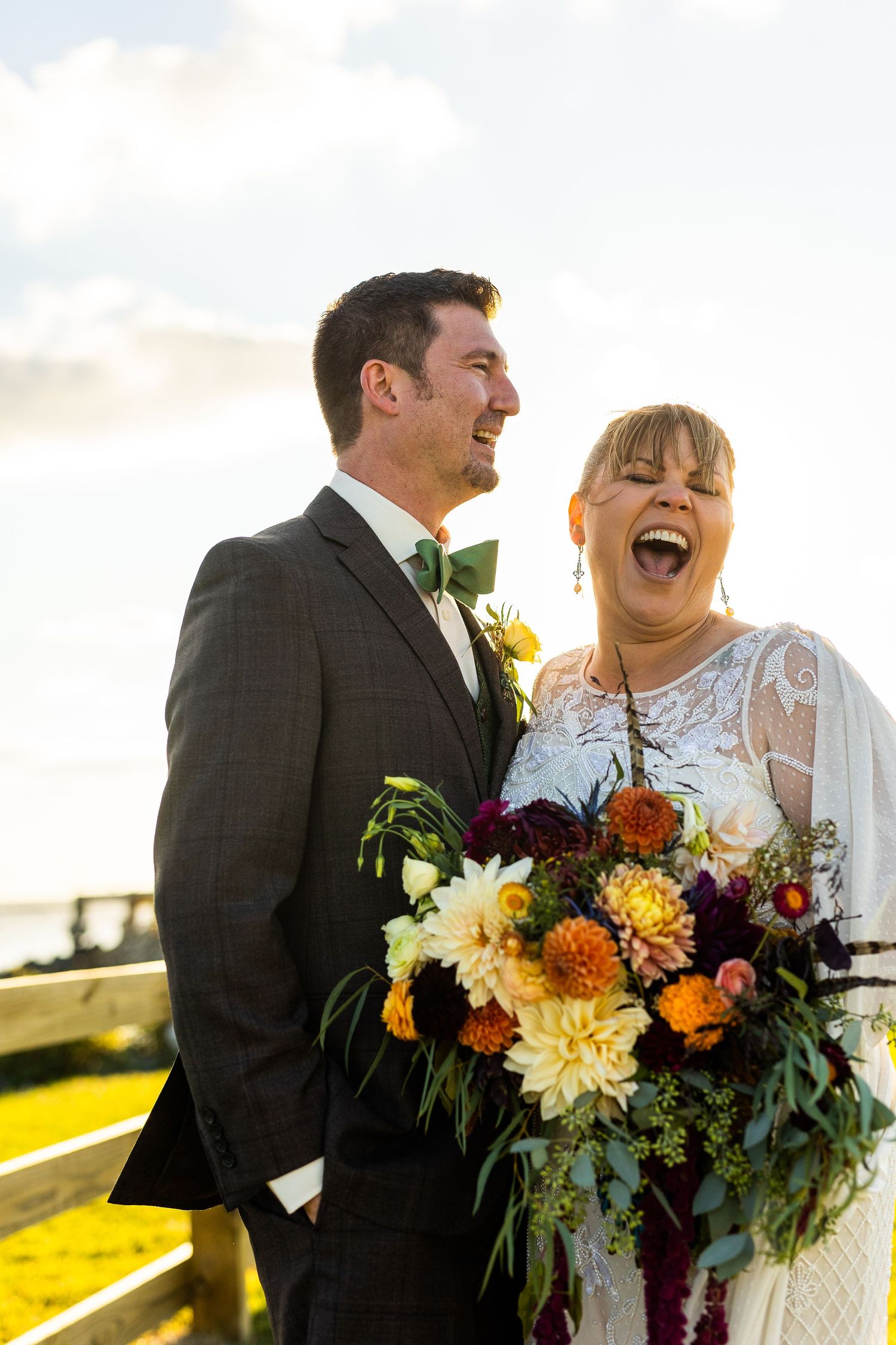 Bride laughing joyfully with groom holding vibrant autumn bouquet at golden hour.