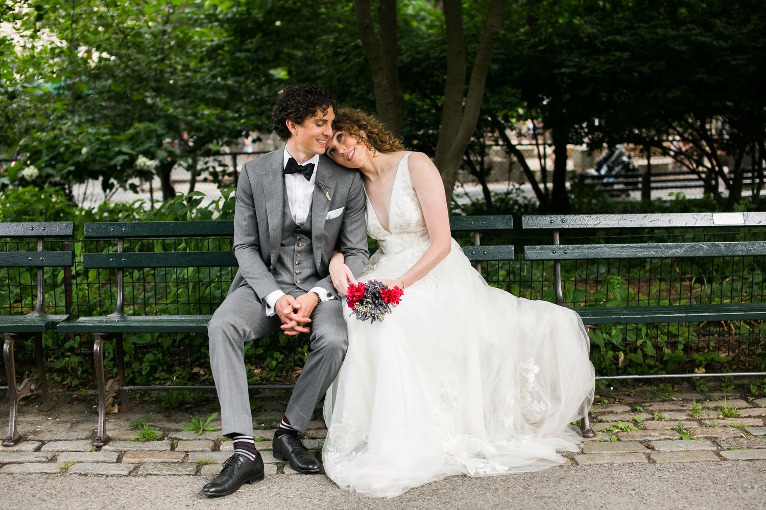 Bride and groom sitting on a park bench, sharing a tender moment in wedding attire with a red bouquet.