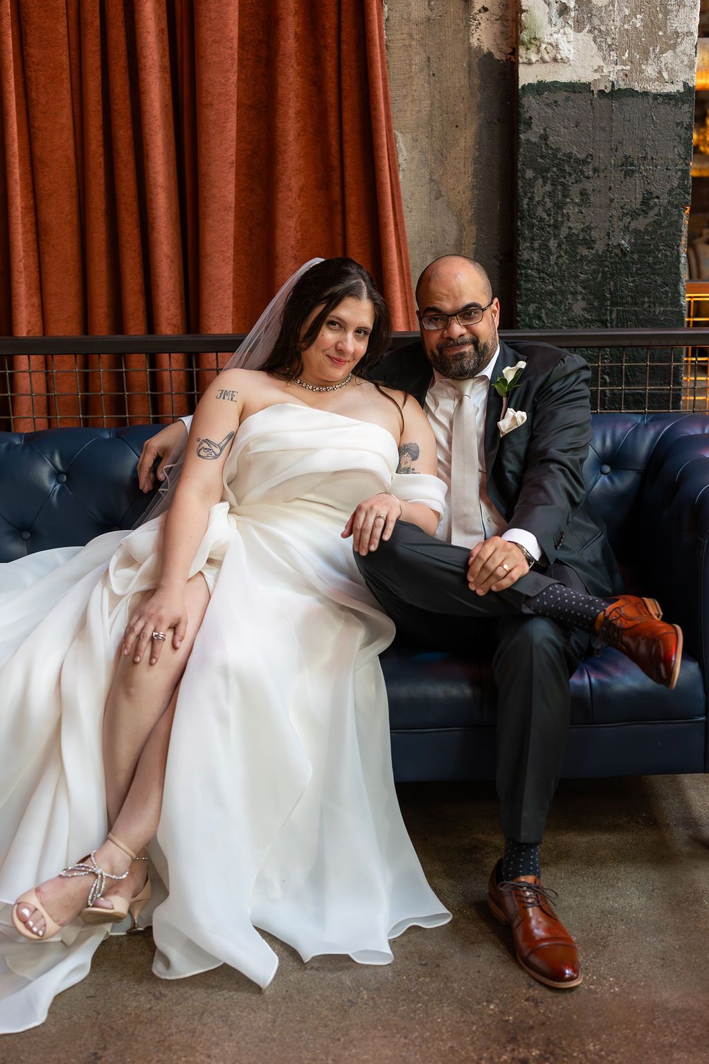 Bride in strapless white gown and groom in dark suit smiling together on blue leather couch at wedding.