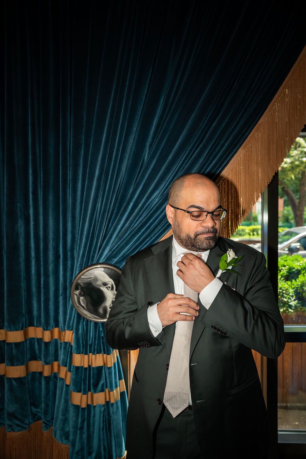 Dapper groom in dark suit adjusting his tie before velvet blue curtains with a dog portrait nearby.