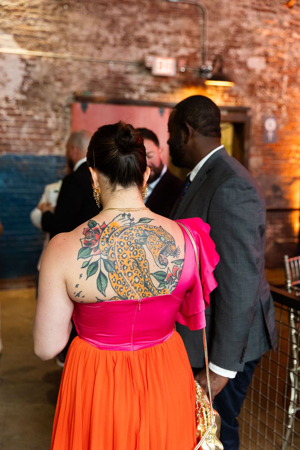 Woman in orange and pink dress with colorful peacock tattoo on her back at an indoor event.