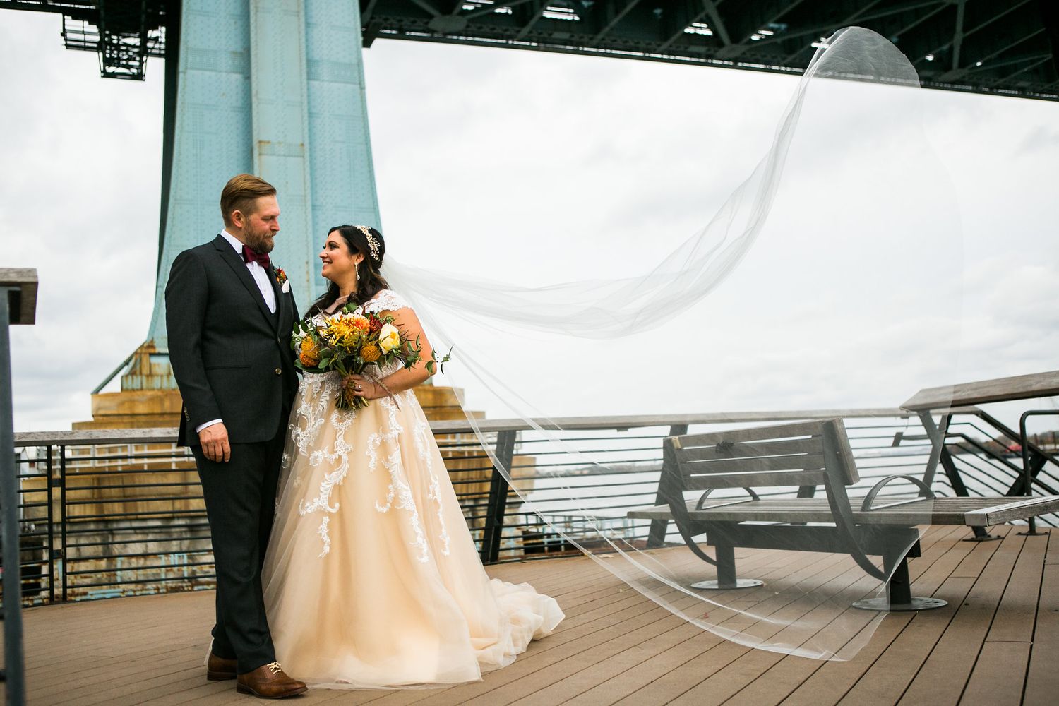 Bride and groom stand together on deck as wedding veil billows dramatically in the wind.