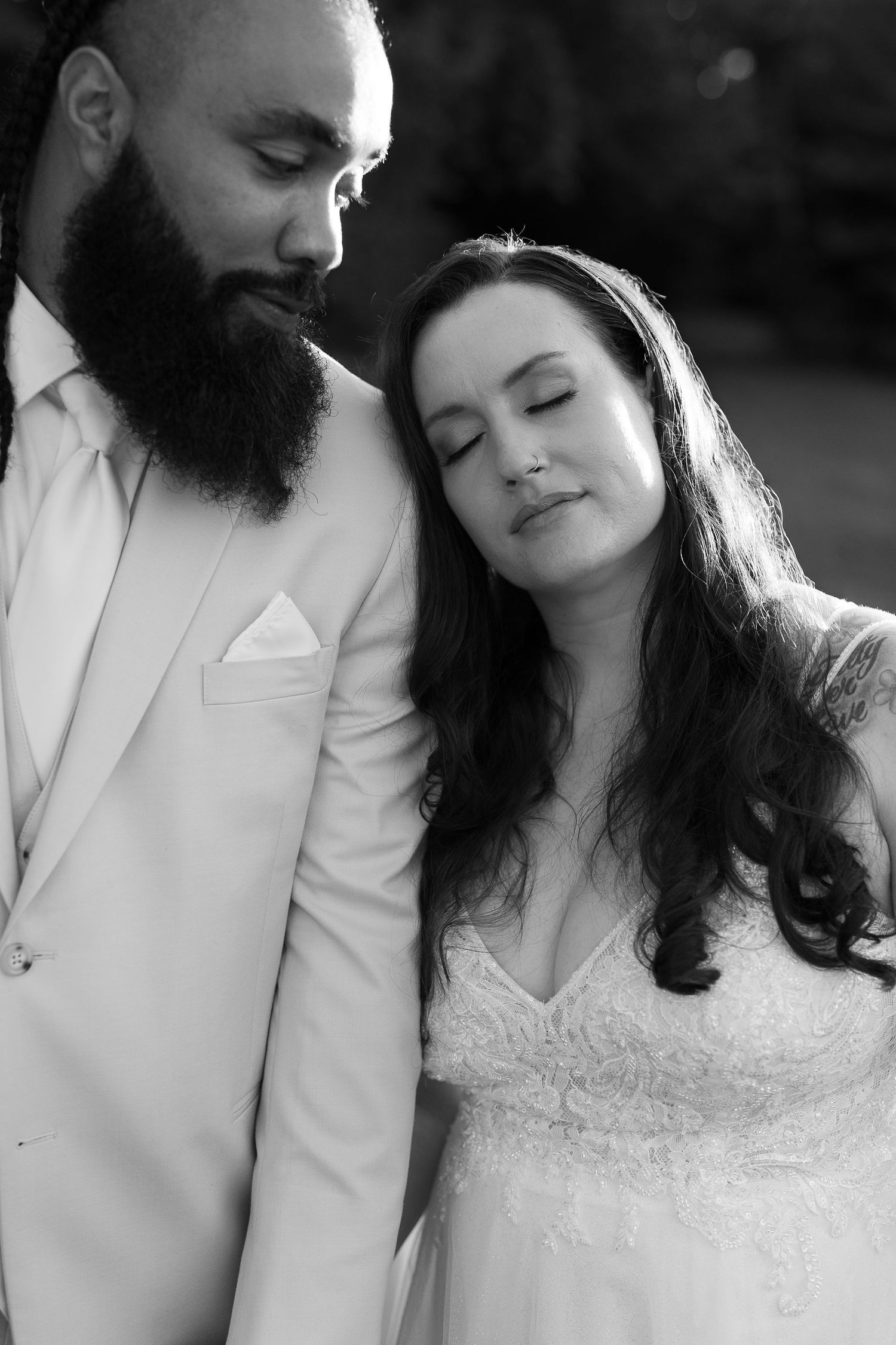 Black and white portrait of a bearded groom in a light suit with bride in lace wedding dress leaning on his shoulder.
