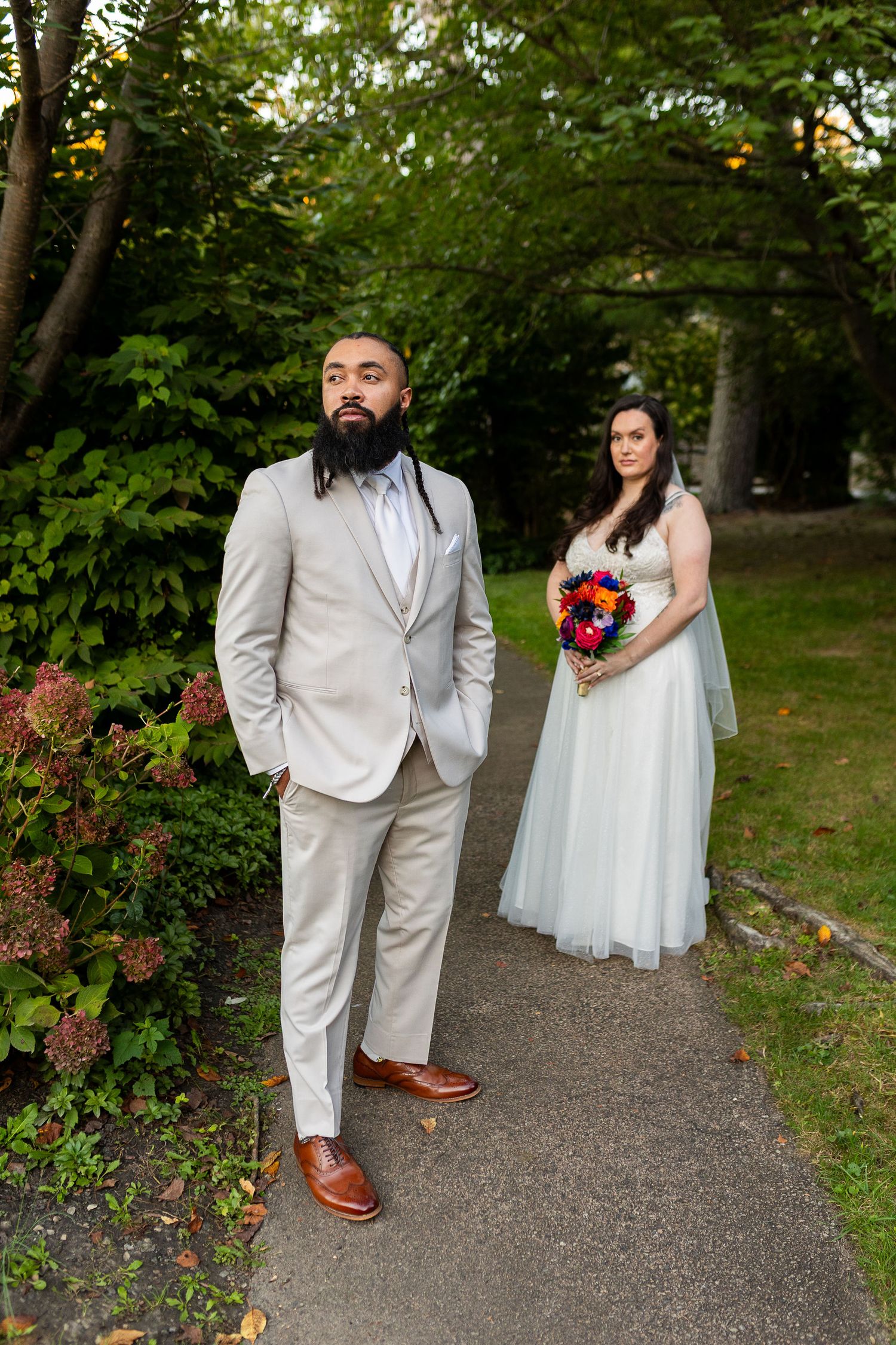 Groom in gray suit and bride in white wedding gown pose on garden pathway at sunset.