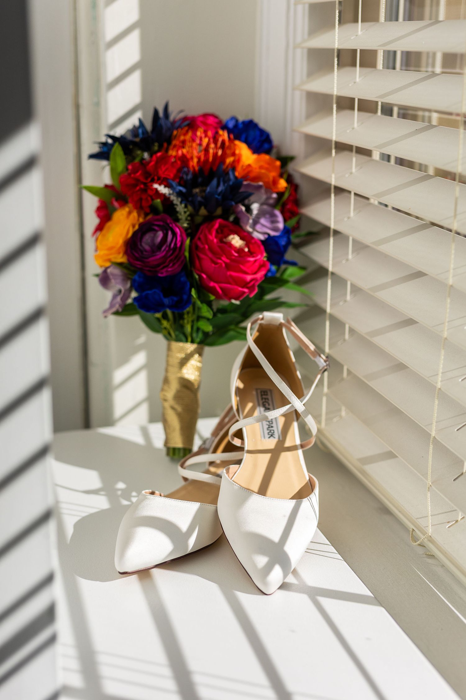 White bridal heels and vibrant colorful bouquet displayed on a windowsill with soft sunlight.