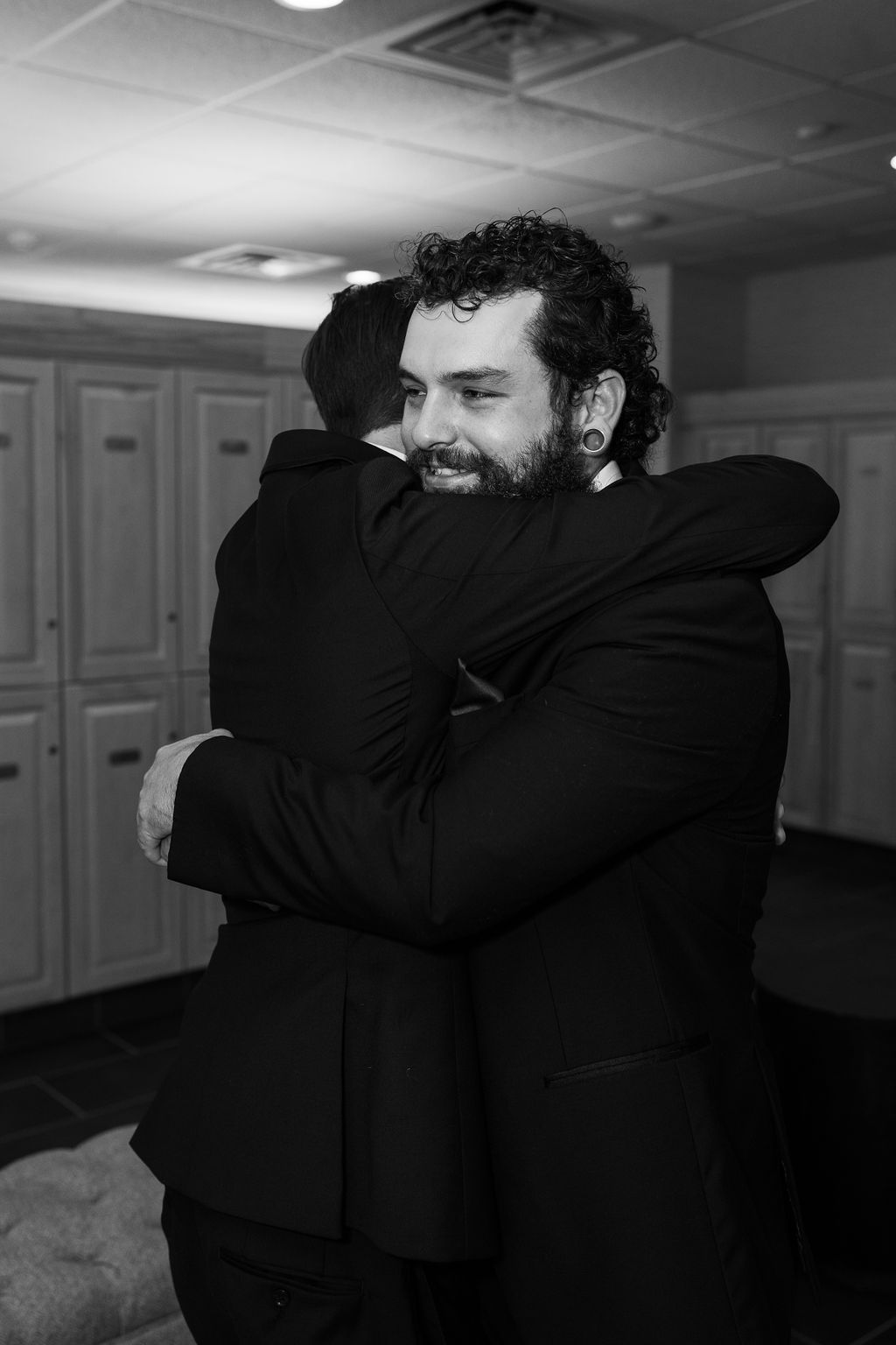 Black and white photo of two men sharing a warm embrace in a locker room before a wedding.