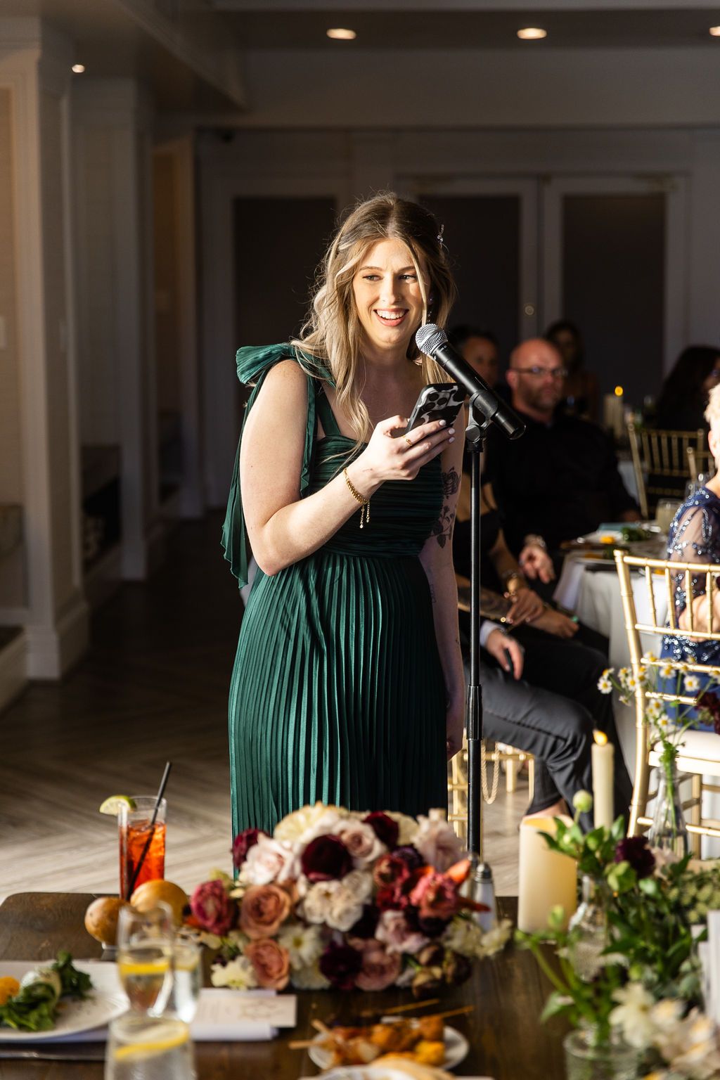 Bridesmaid in emerald green pleated dress delivers speech at microphone during wedding reception.
