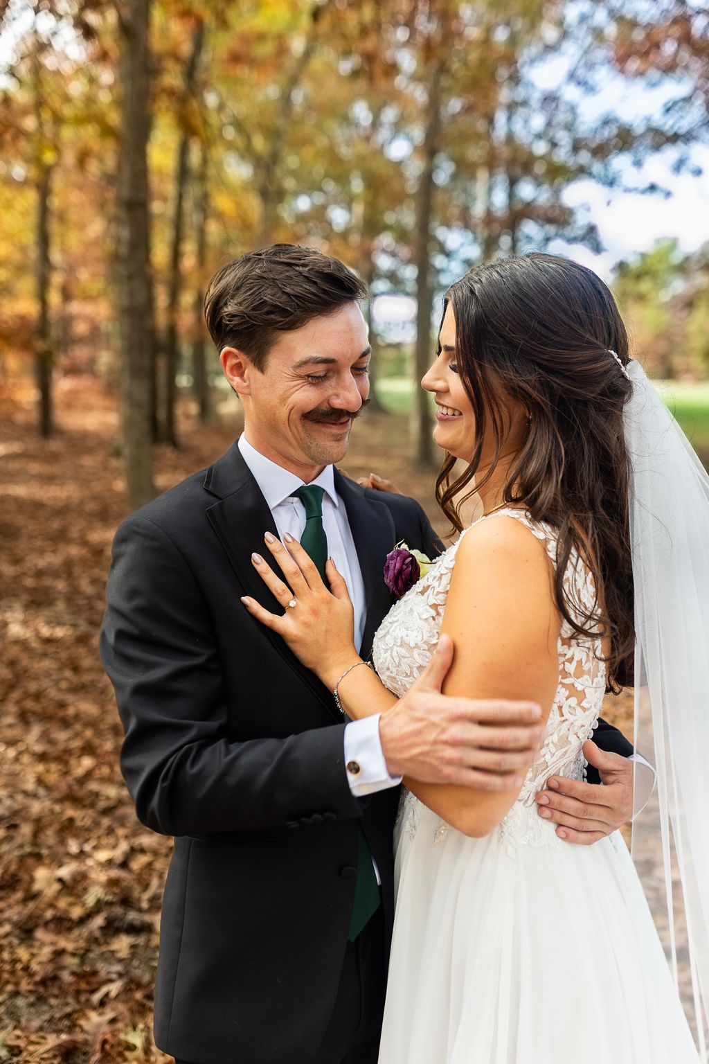 Smiling bride and mustachioed groom embrace warmly among vibrant fall trees in wedding attire.