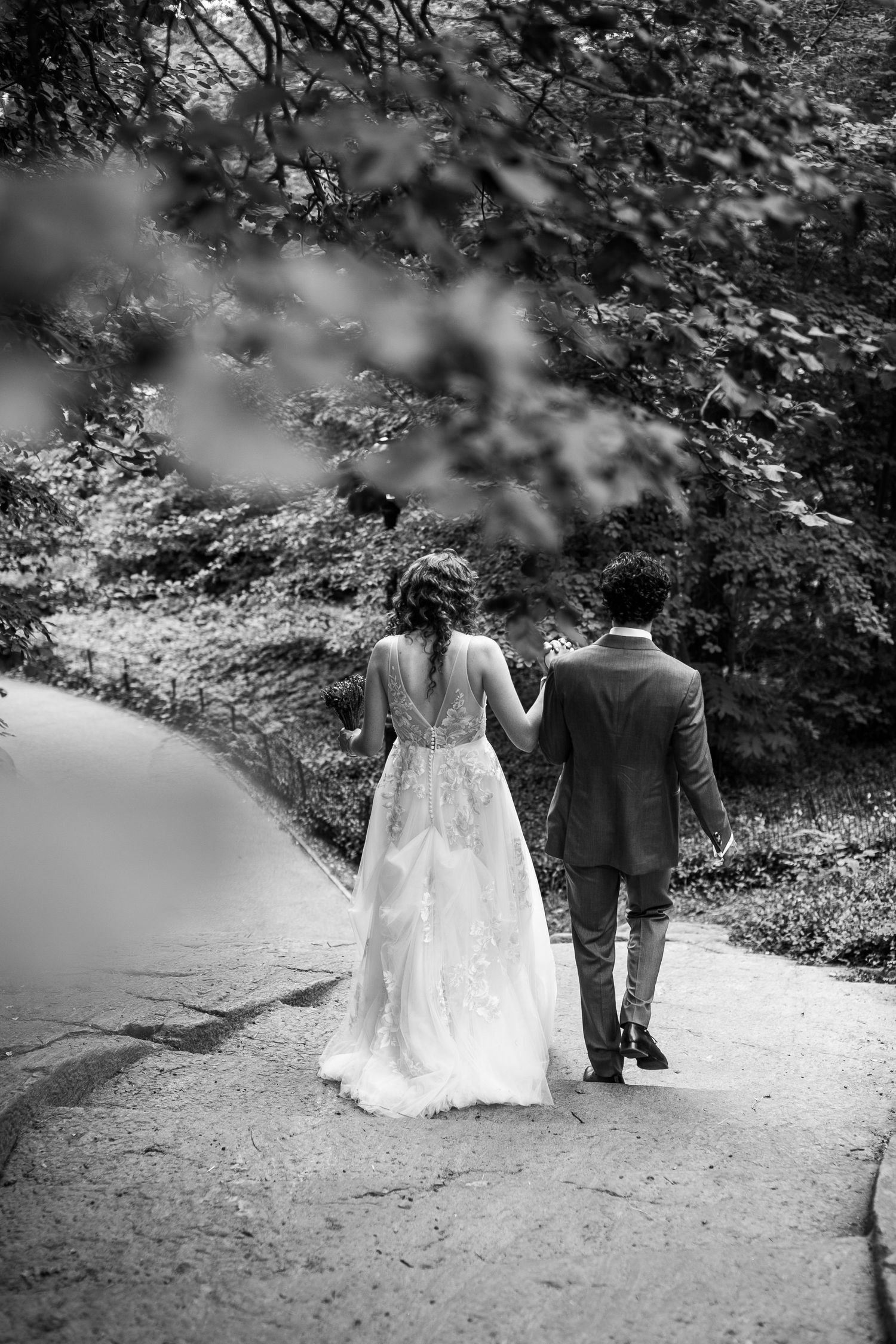 Black and white photo of bride and groom walking hand in hand along a wooded path.