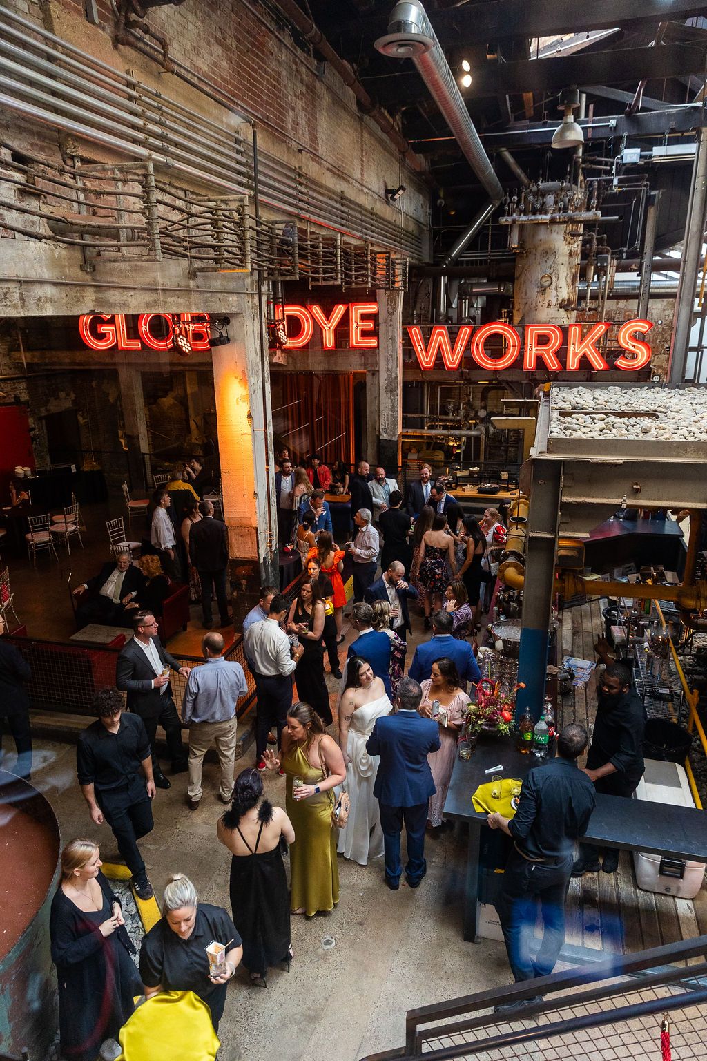 Wedding guests mingle under illuminated Globe Dye Works neon sign in a rustic industrial event space.