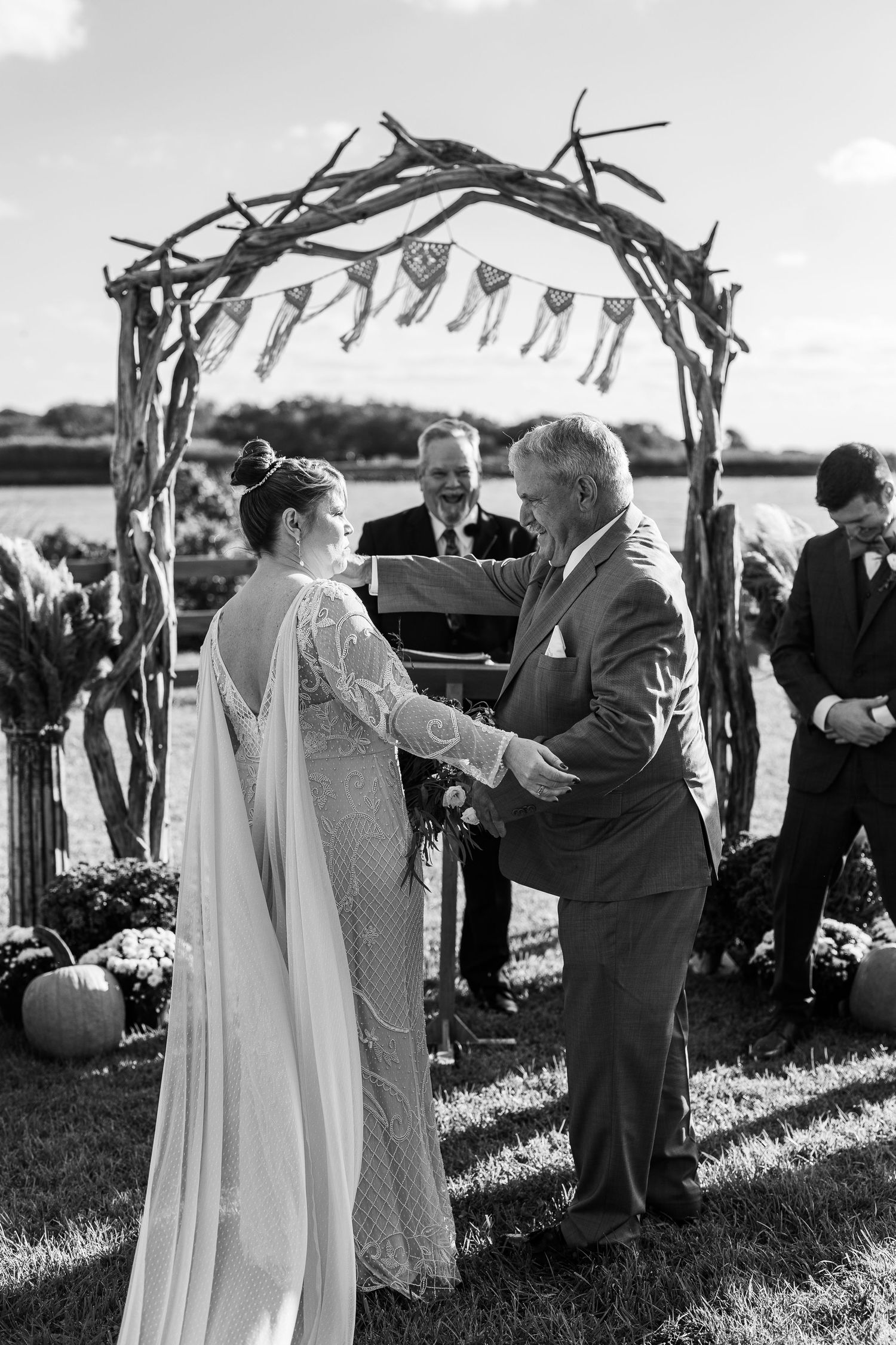 Black and white photo of bride and groom exchanging vows under rustic driftwood arch at outdoor wedding.