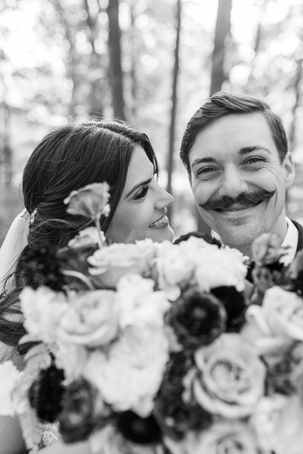 Black and white portrait of laughing bride and mustachioed groom holding floral bouquet outdoors.