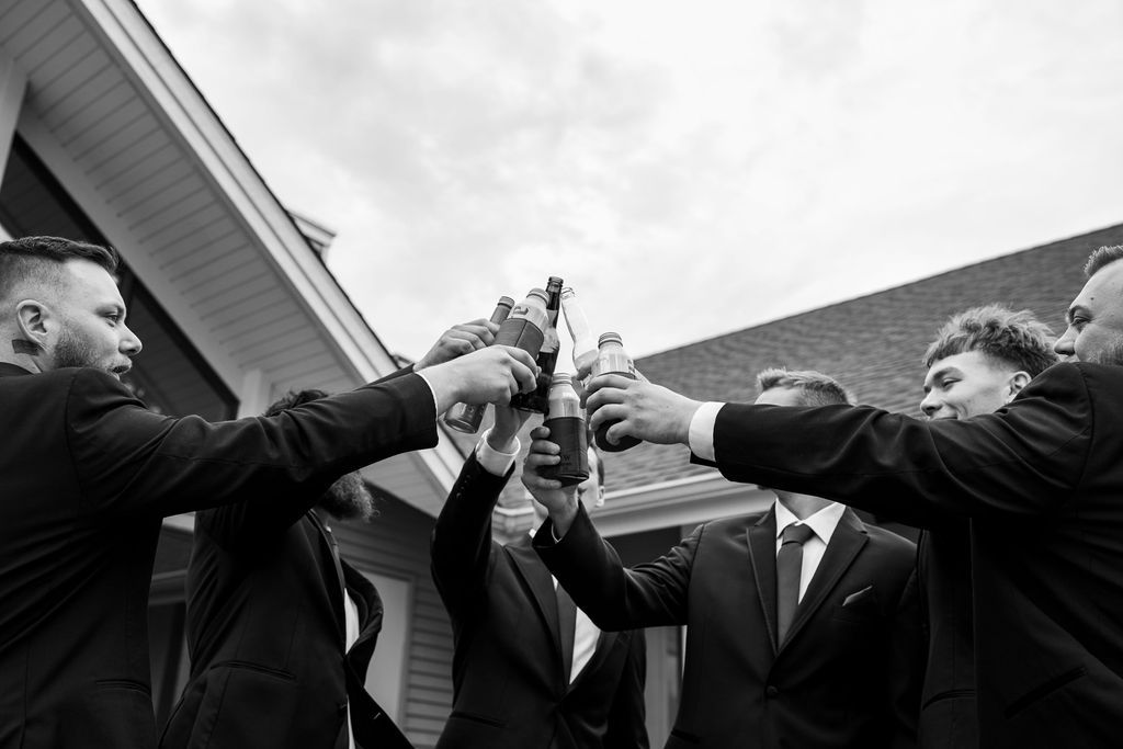 Groomsmen in black suits toasting with beer bottles in black and white wedding celebration photo.