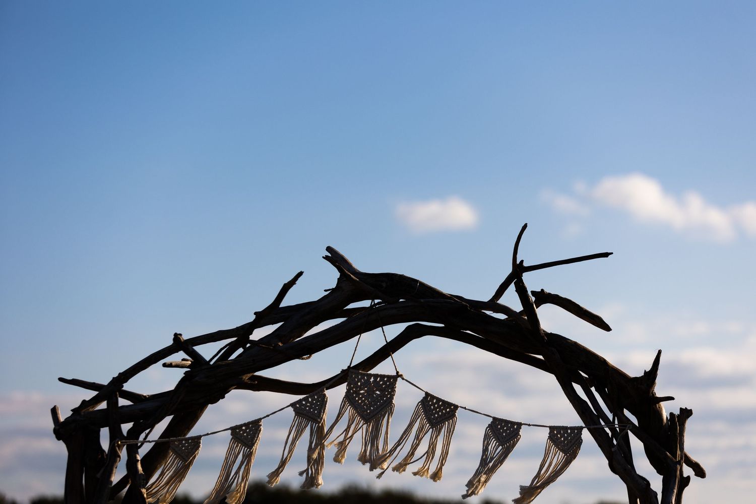 Rustic driftwood wedding arch with macrame bunting silhouetted against a bright blue sky.