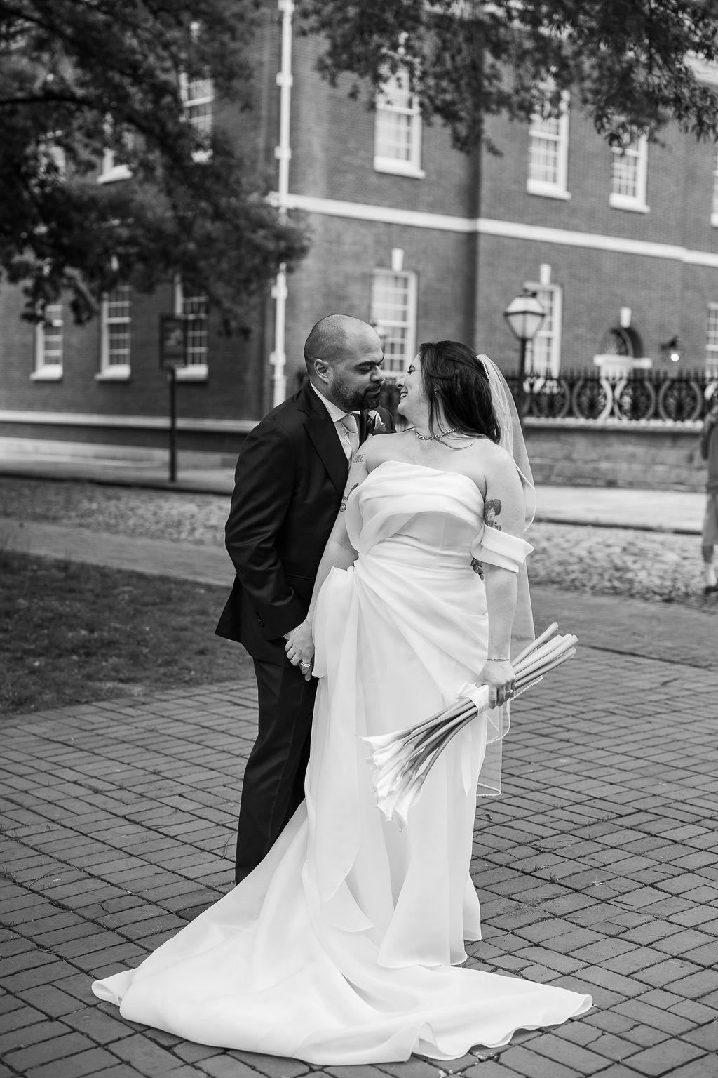 Black and white romantic photo of bride and groom sharing a kiss on brick path with historic building backdrop.