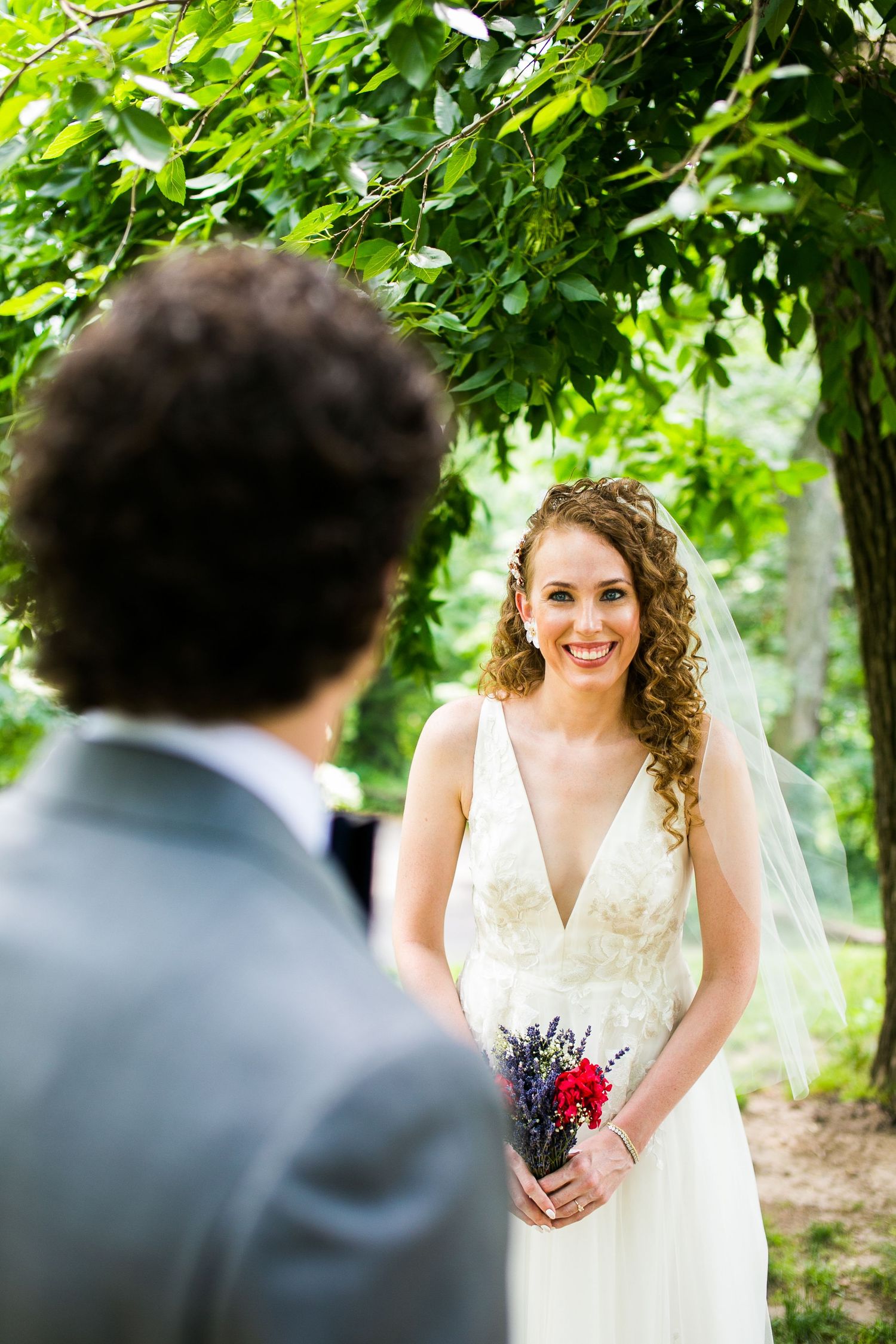Radiant bride in white V-neck gown and veil smiles at groom during outdoor first look ceremony.