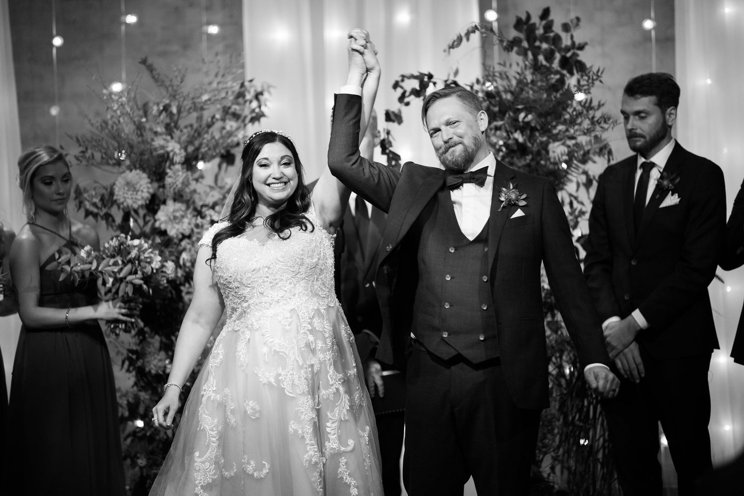 Groom raises hand in celebration beside bride in lace gown at wedding reception.