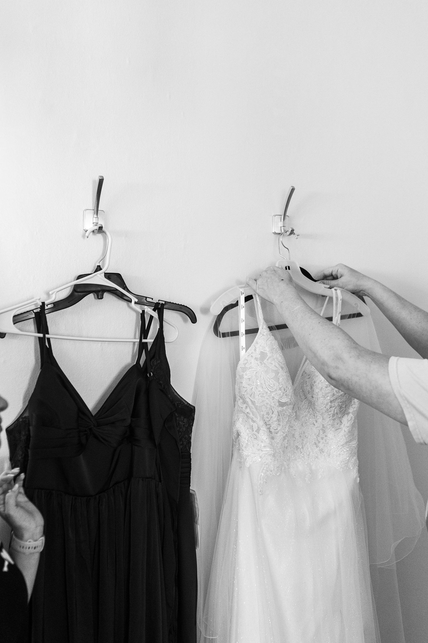 Black and white photo of a white wedding dress and black bridesmaid dress hanging on wall hooks.