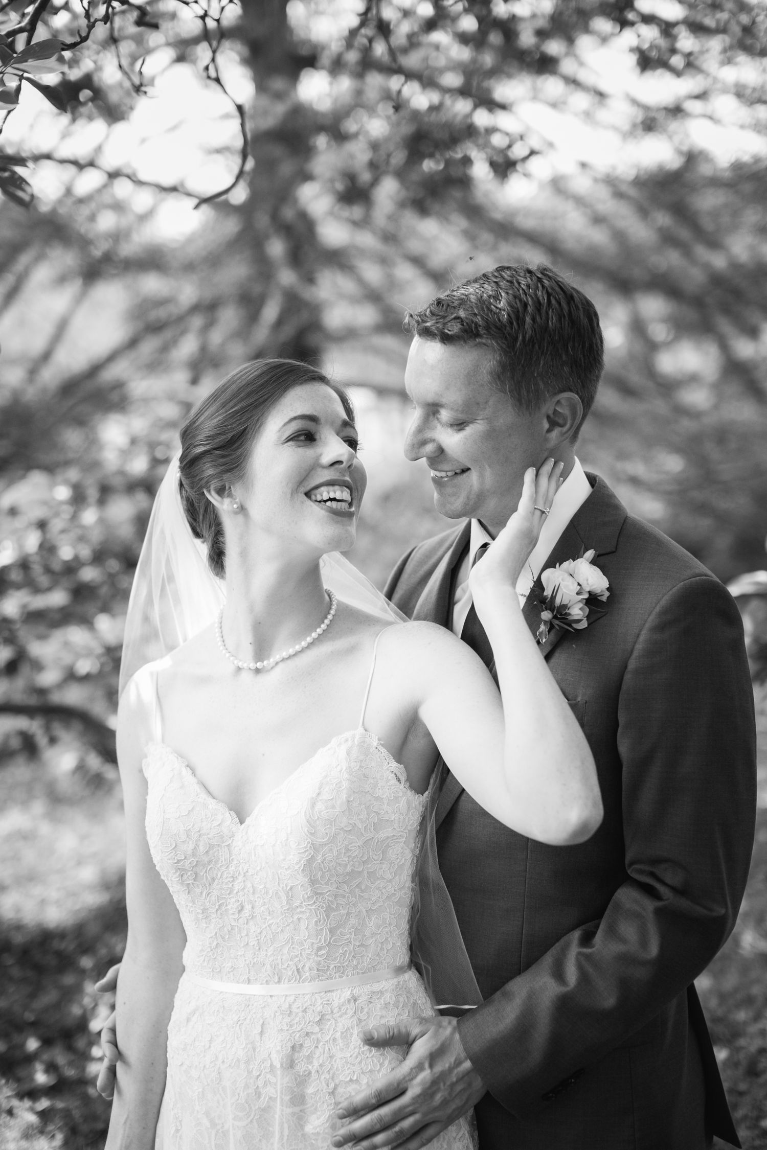 Black and white wedding portrait of a joyful bride touching her groom's face under a canopy of trees.