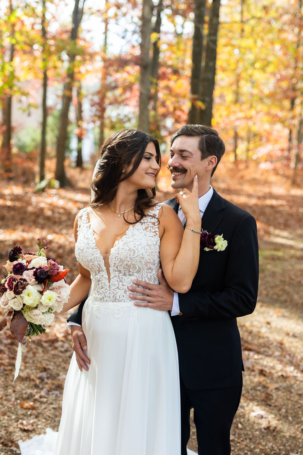 Bride in lace wedding dress gazing at groom with mustache holding bouquet in vibrant autumn woodland.