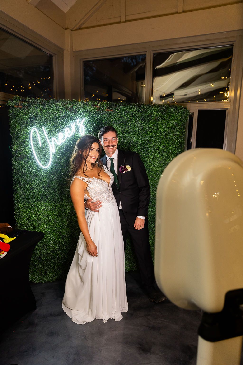 Bride and groom smile together at a wedding photo booth featuring a lush green hedge and neon Cheers sign.