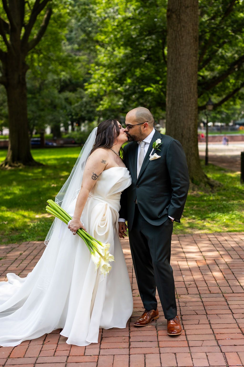 Bride and groom share a romantic kiss while holding hands on a brick path in a lush green park.